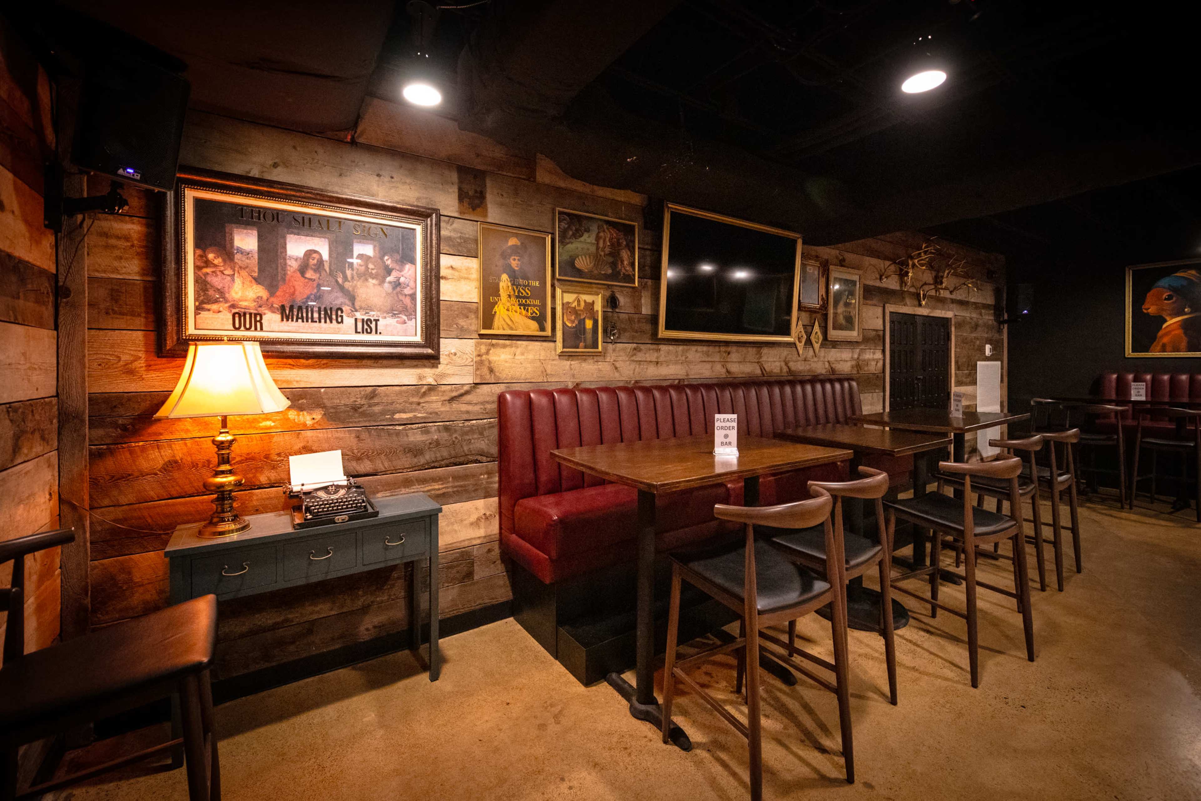 The image shows a rustic dining area with wooden walls, a red leather booth, a table with chairs, and framed artwork.