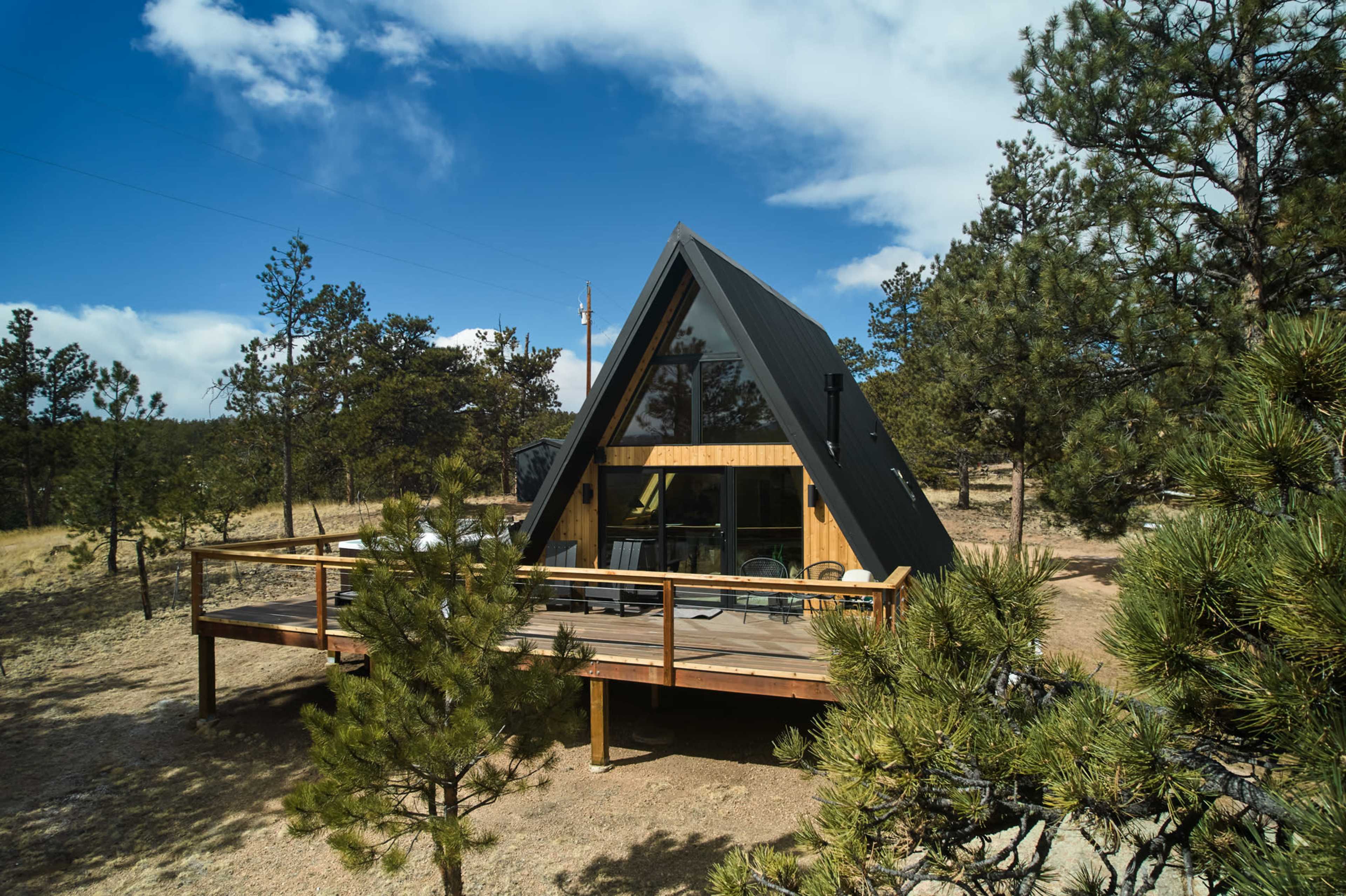A modern A-frame cabin with a wooden deck is surrounded by pine trees under a blue sky with scattered clouds.