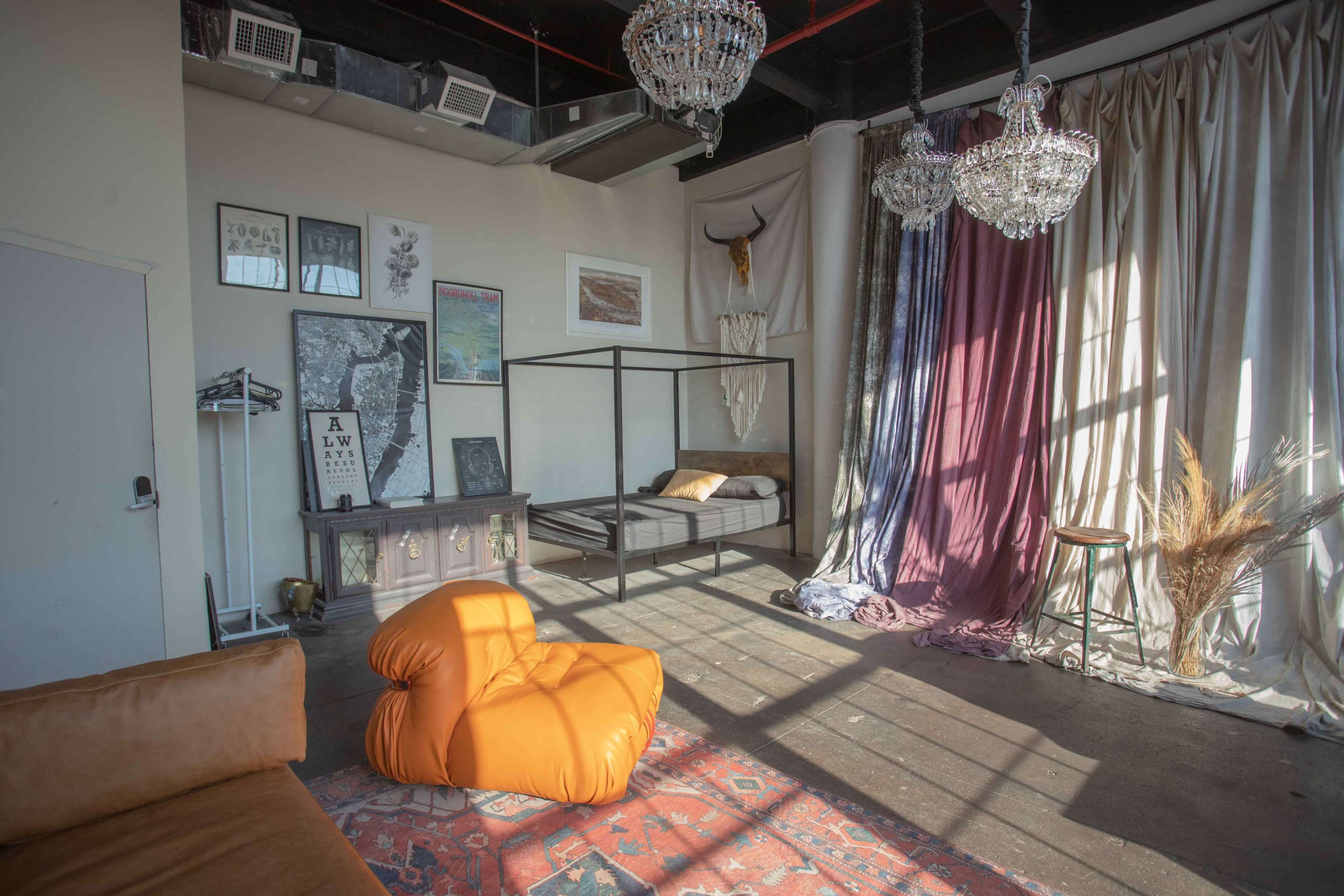 A spacious room featuring a four-poster bed, an orange bean bag chair, a decorative chest, and large windows casting shadows on the floor.