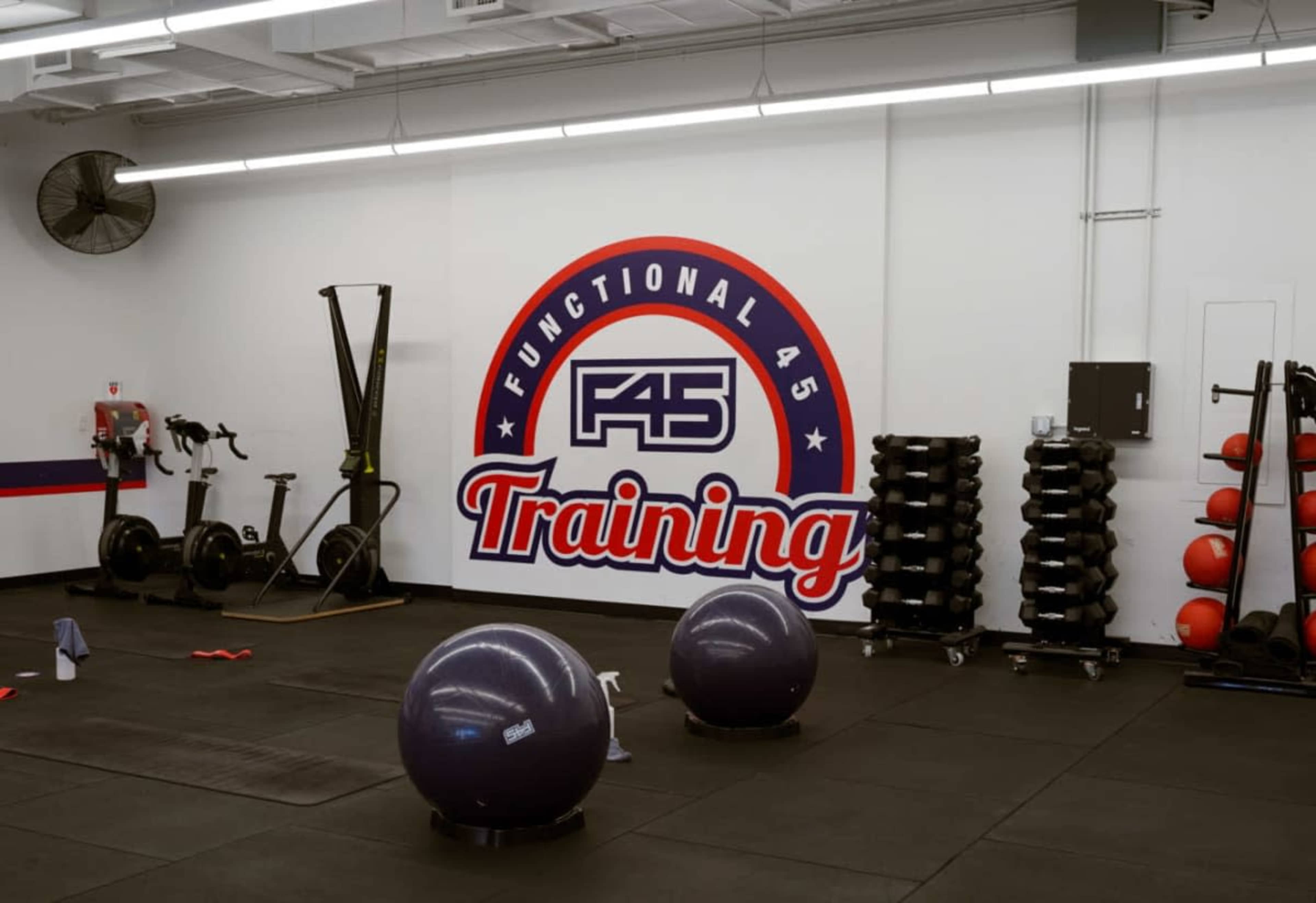 The image shows a gym interior with exercise equipment, including kettlebells, exercise balls, and cardio machines, along with a prominent wall logo for F45 Training.