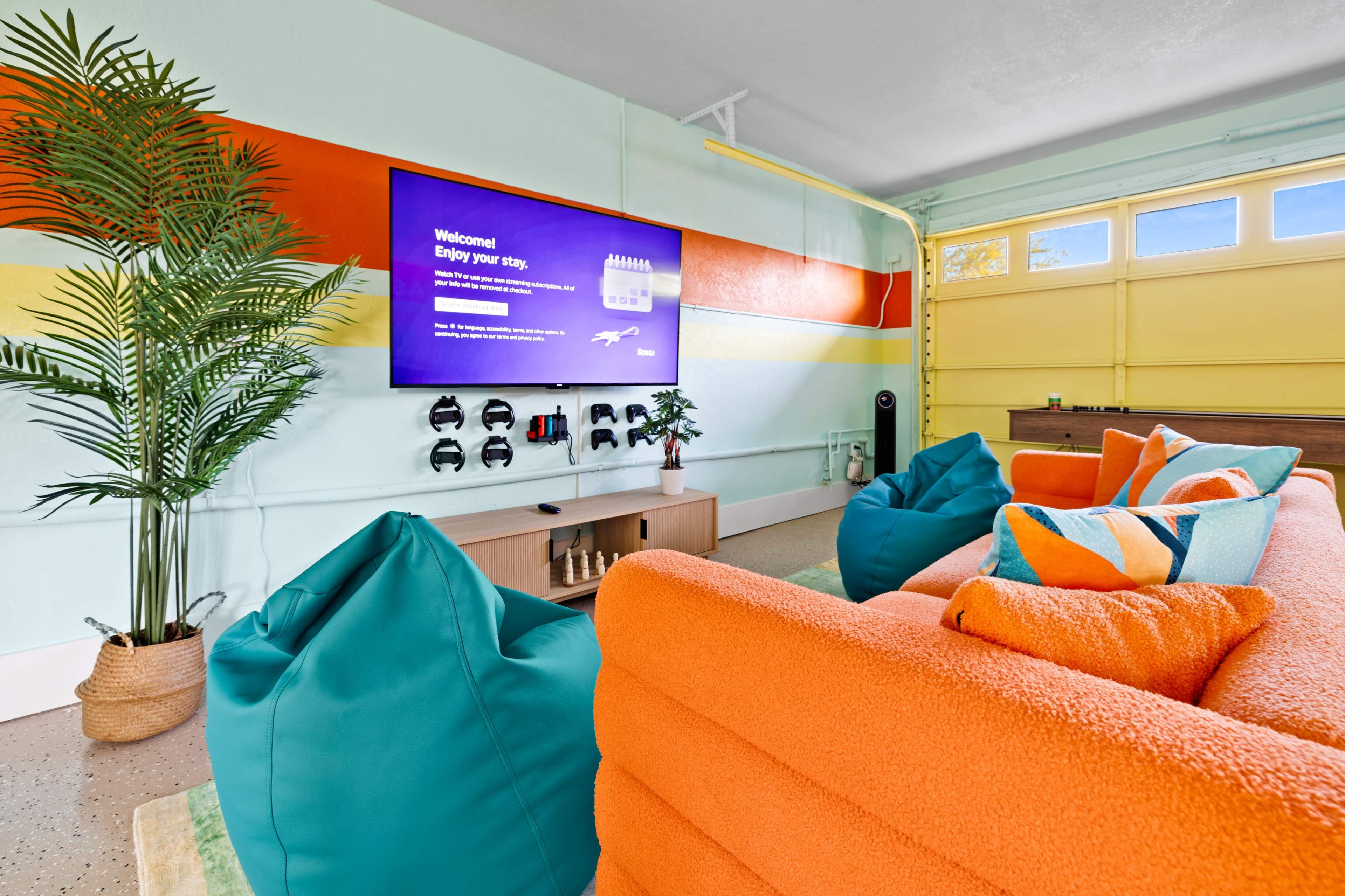 The image shows a bright, modern living space with a large television mounted on the wall and a colorful sofa positioned in front of it, surrounded by bean bags and a potted plant.