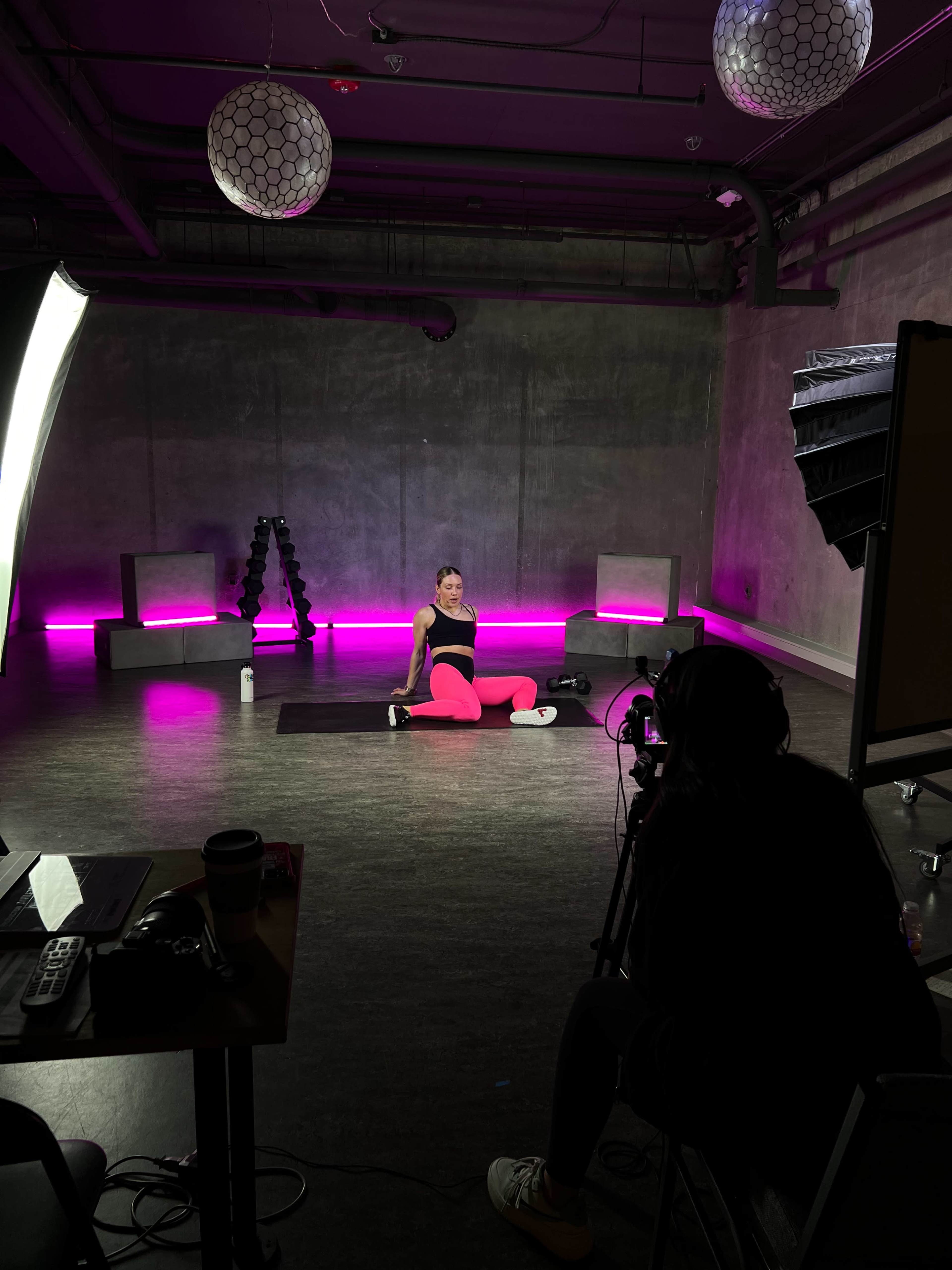 A person is sitting on a yoga mat in a dimly lit studio with pink lighting, while another individual is seated at a table, focused on operating a camera.