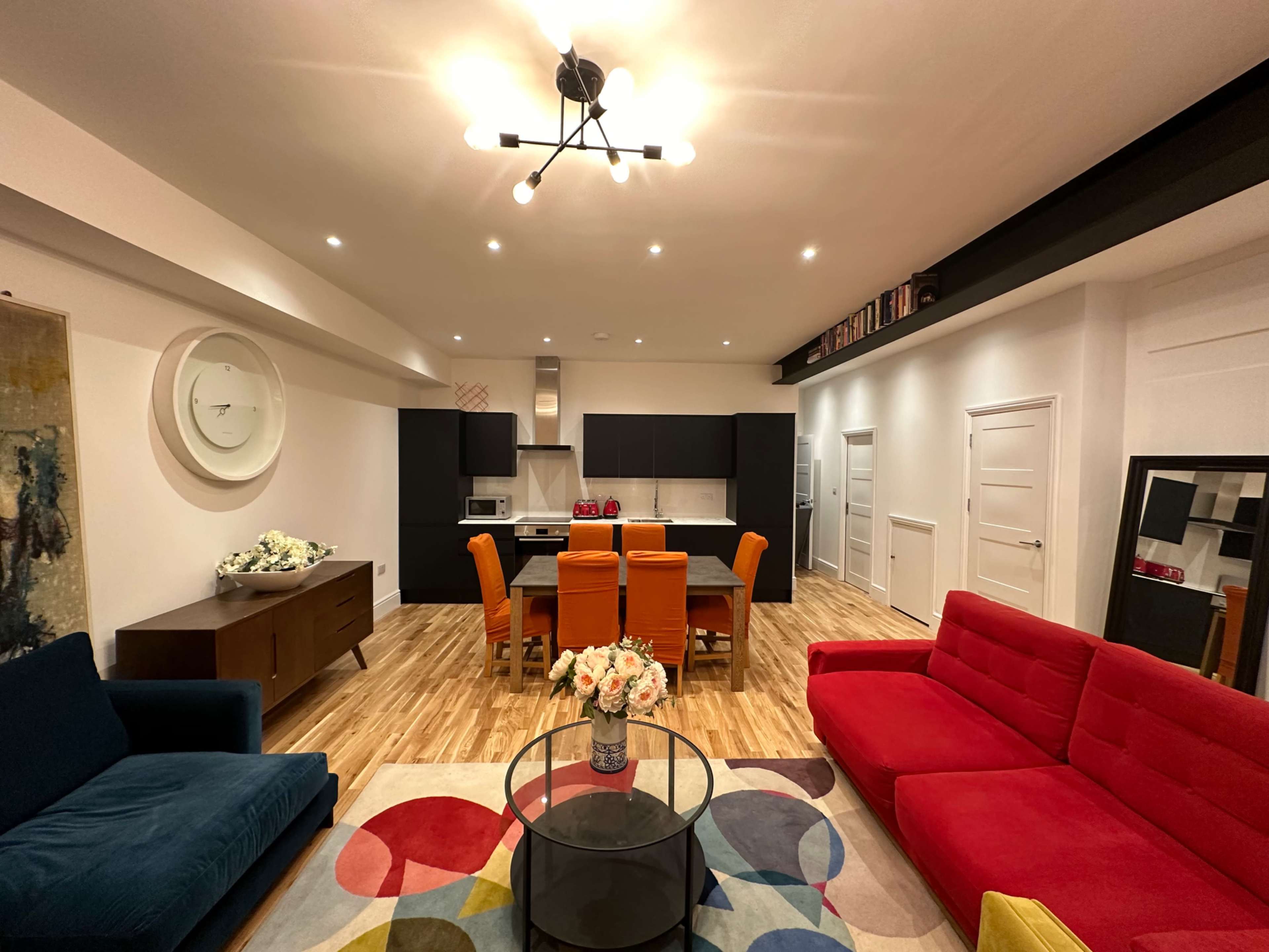 A modern living space featuring a red sofa, a blue couch, a circular coffee table, a dining area with orange chairs, and a kitchen in the background.