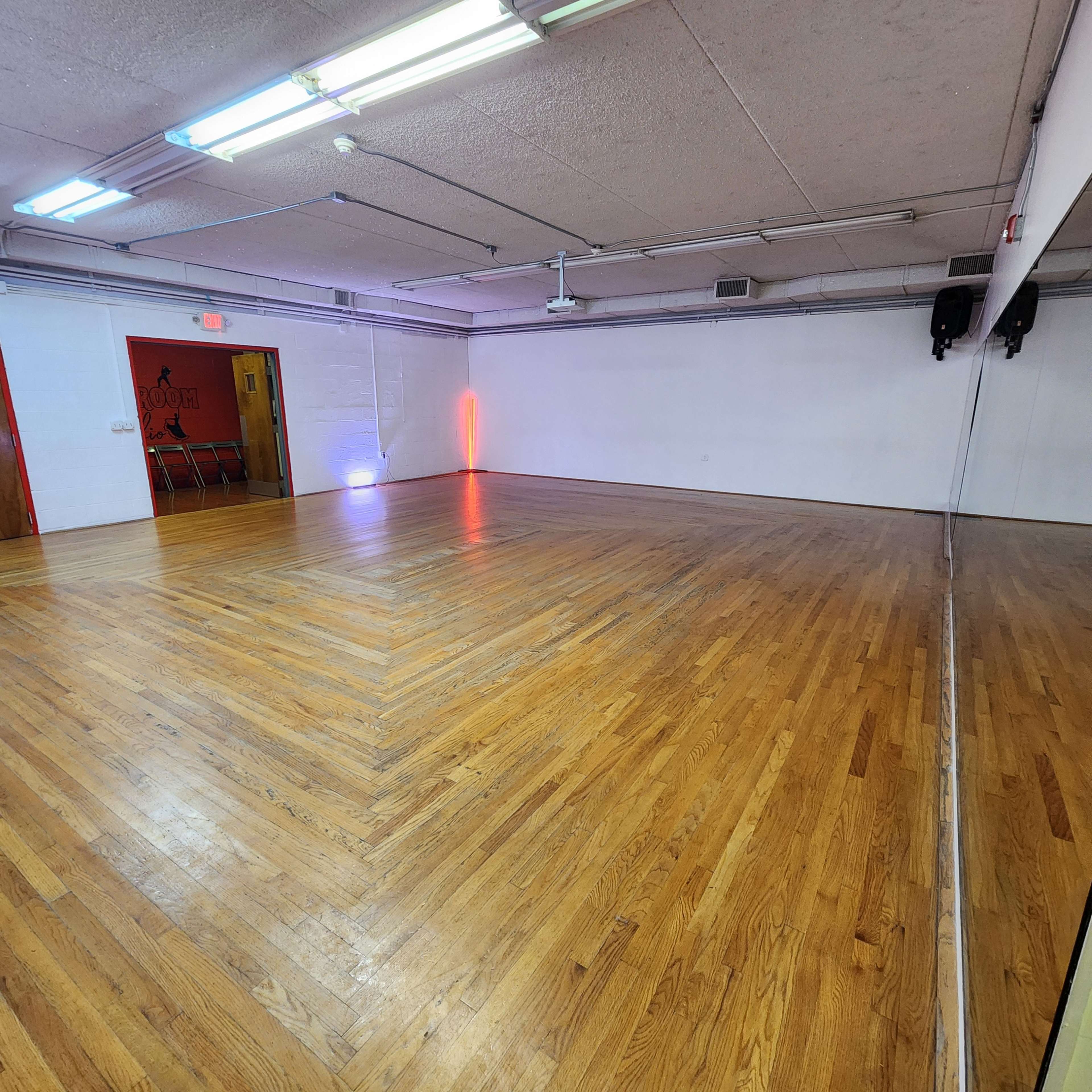 The image shows an empty room with hardwood flooring, featuring a wall of mirrors and an entrance at one end with colored lights illuminating the space.
