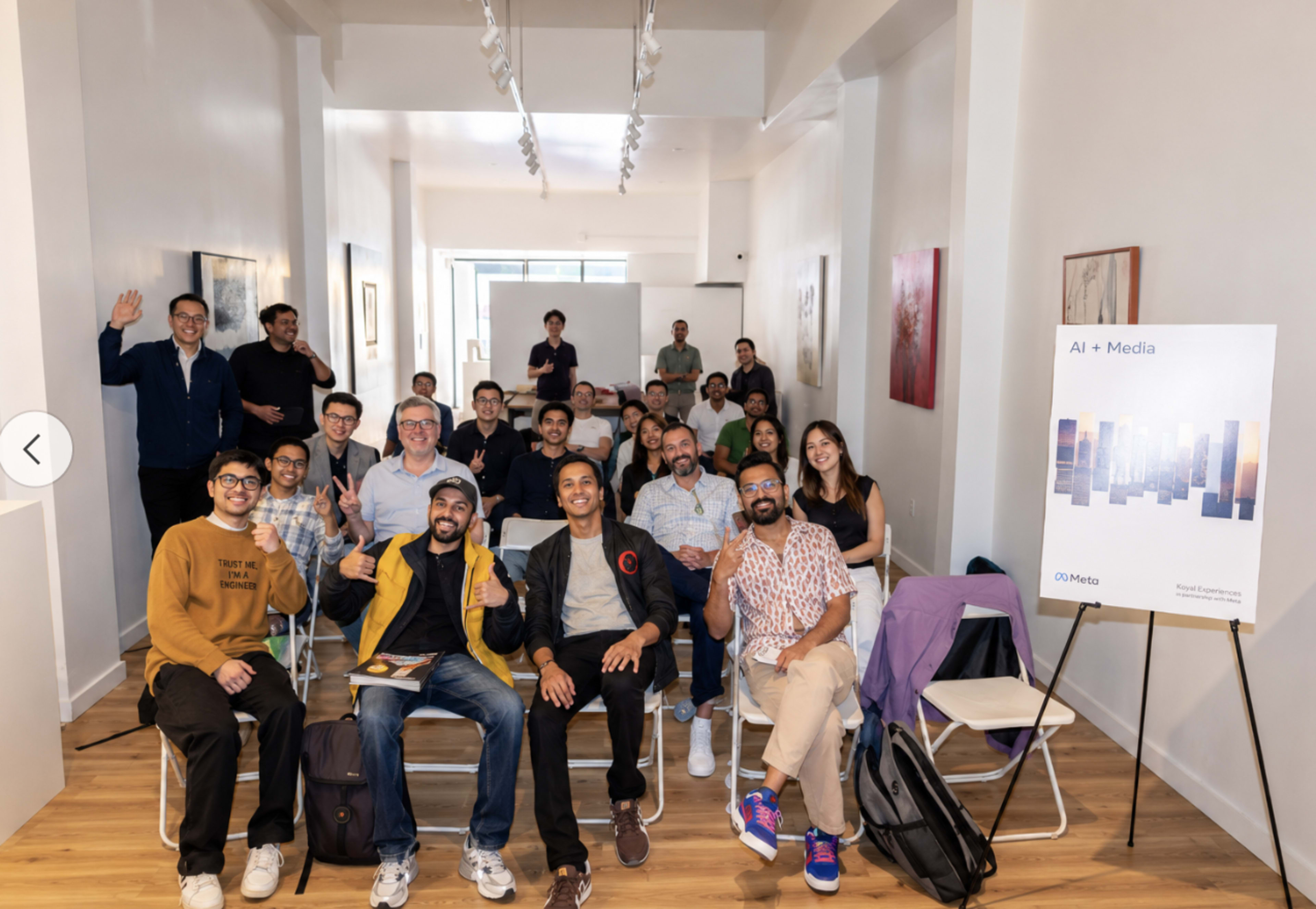 A group of diverse individuals is posing for a photo in a well-lit room, seated and standing around a sign that reads "AI + Media."
