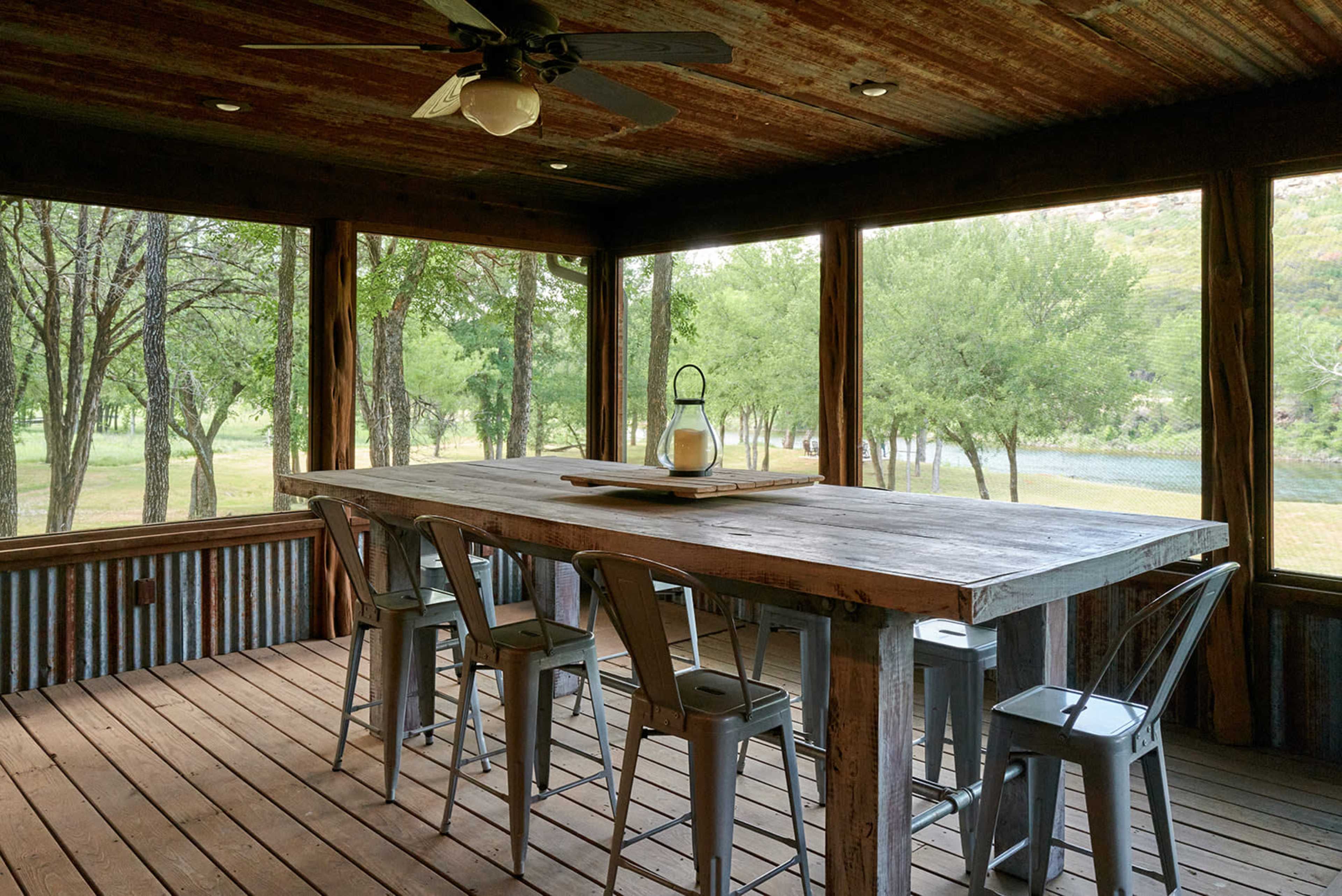 A spacious, rustic dining area features a large wooden table surrounded by metal chairs, with large windows offering views of a green landscape.
