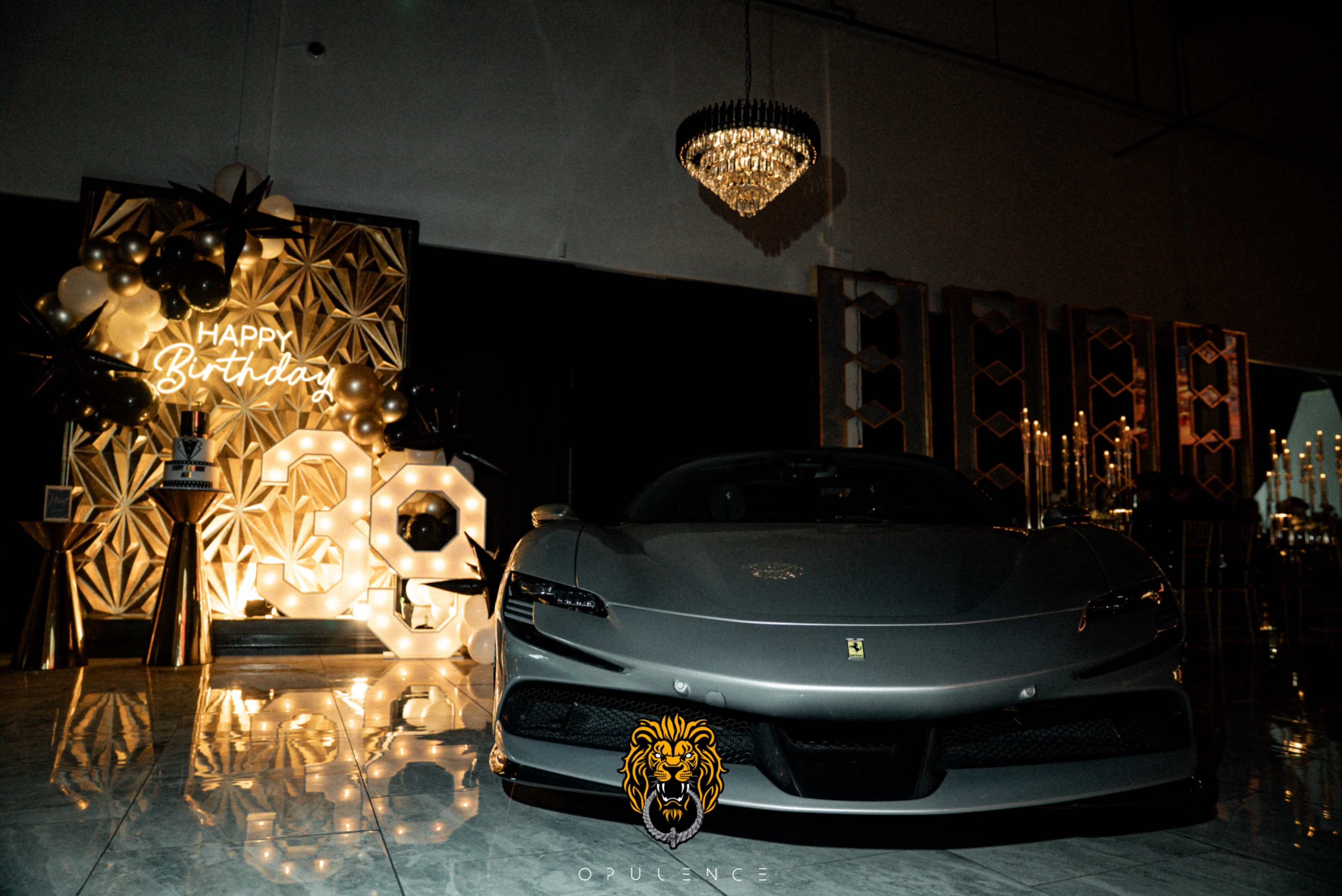 A sleek gray sports car is positioned prominently in front of a festive birthday display featuring balloons and decorative elements.