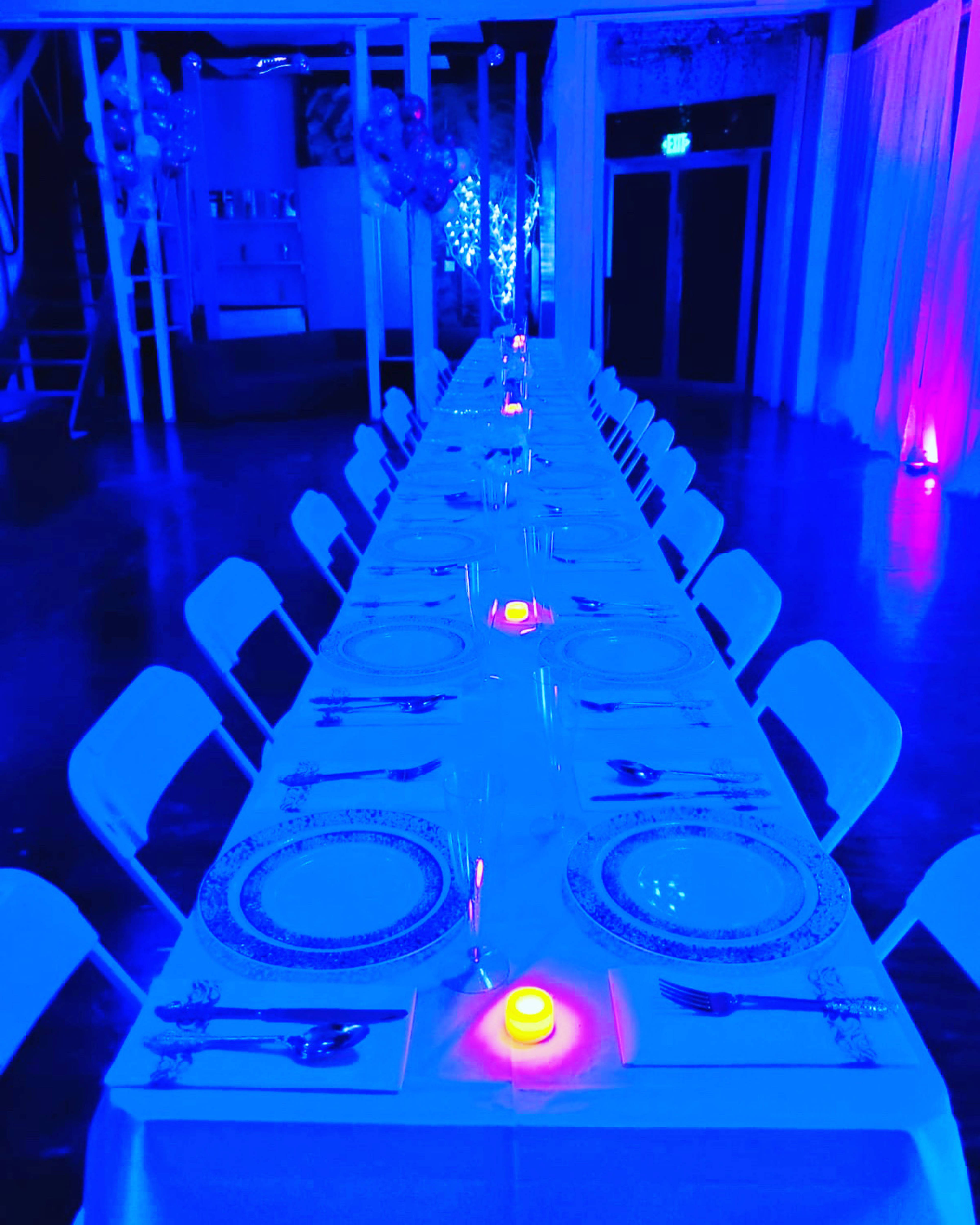 A long banquet table is set with plates, cutlery, and candles under blue lighting in an event space.
