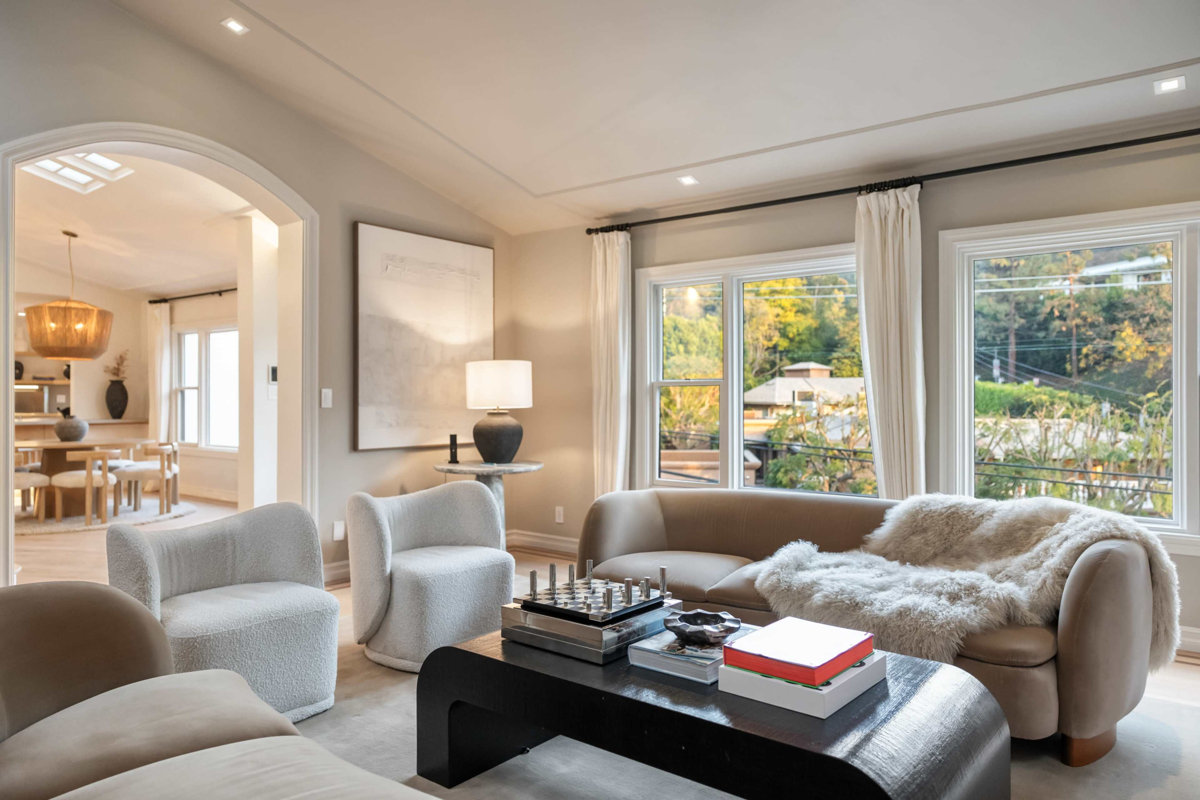 The image shows a modern, well-lit living room with a comfortable sofa, two armchairs, a coffee table with a chess set, and large windows overlooking greenery.