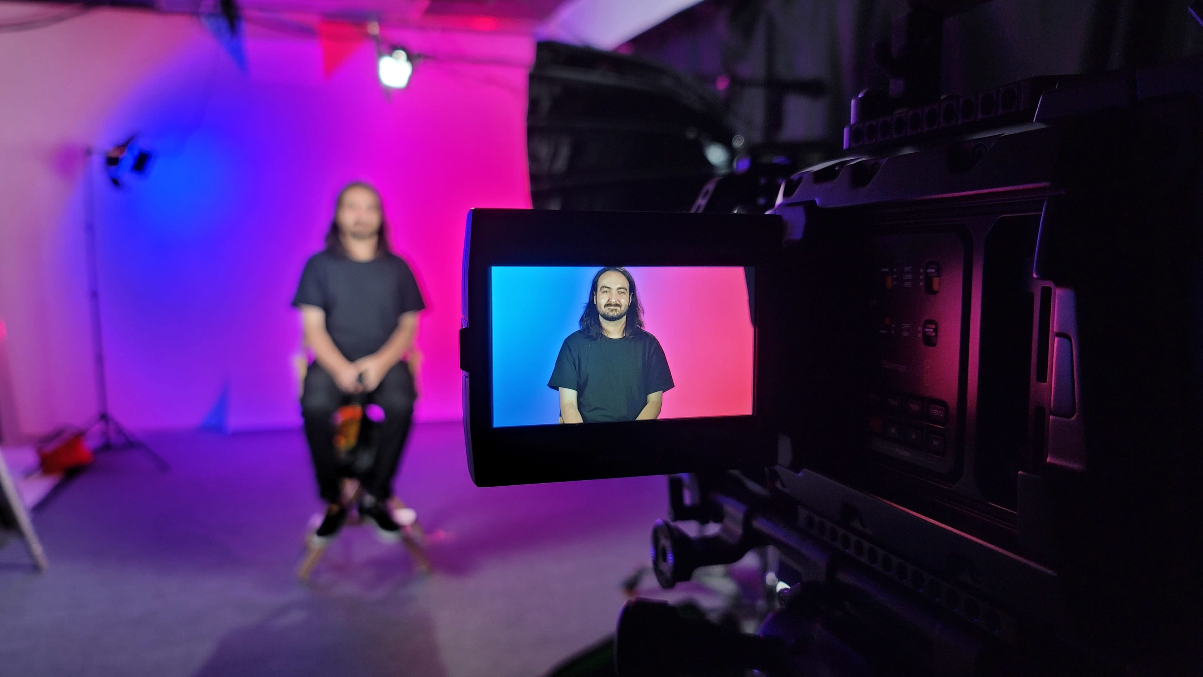 A camera displays a man sitting on a stool in front of a colored backdrop, with lighting effects creating a contrasting pink and blue environment.