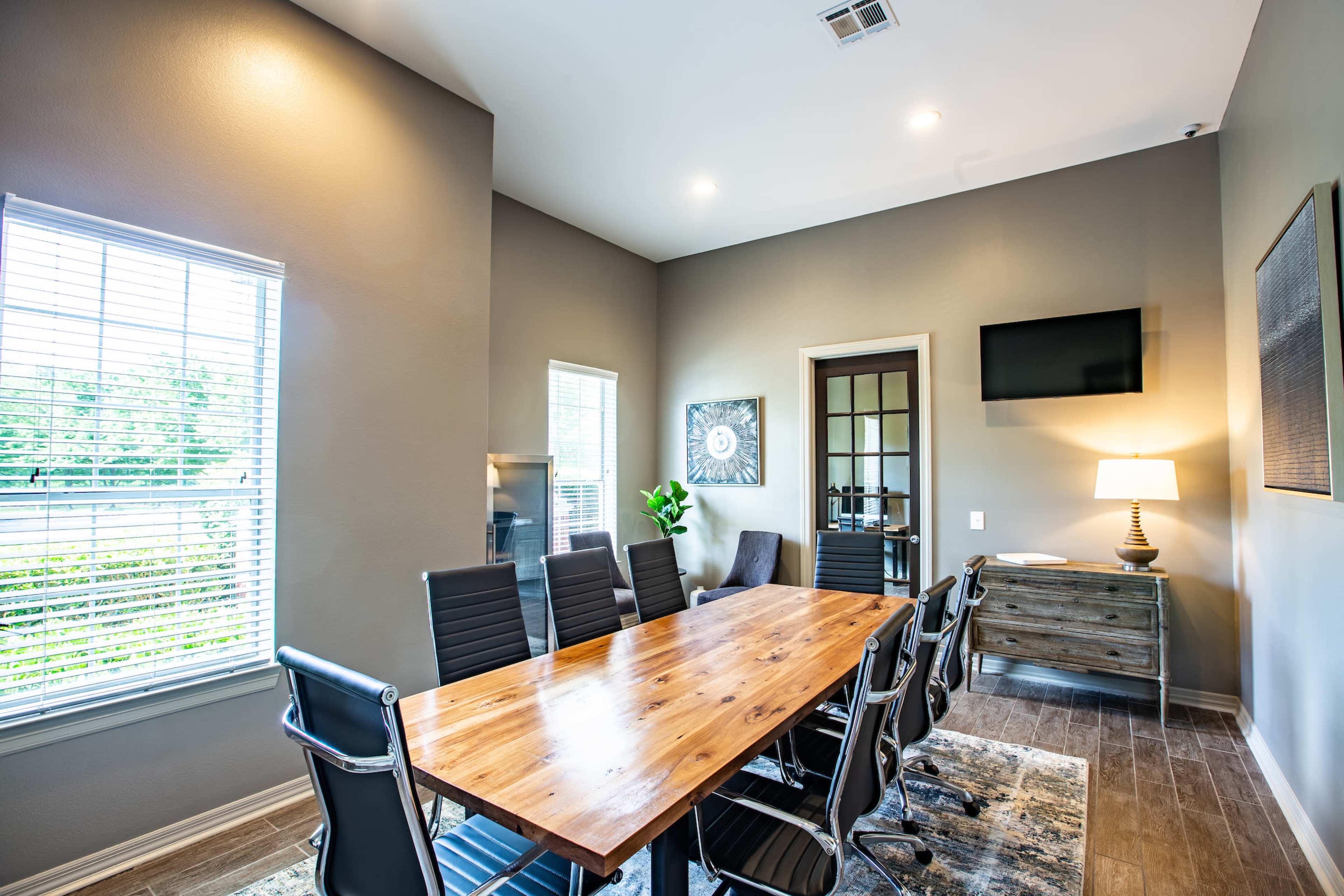A modern conference room features a long wooden table surrounded by black chairs, with large windows and a TV mounted on the wall.