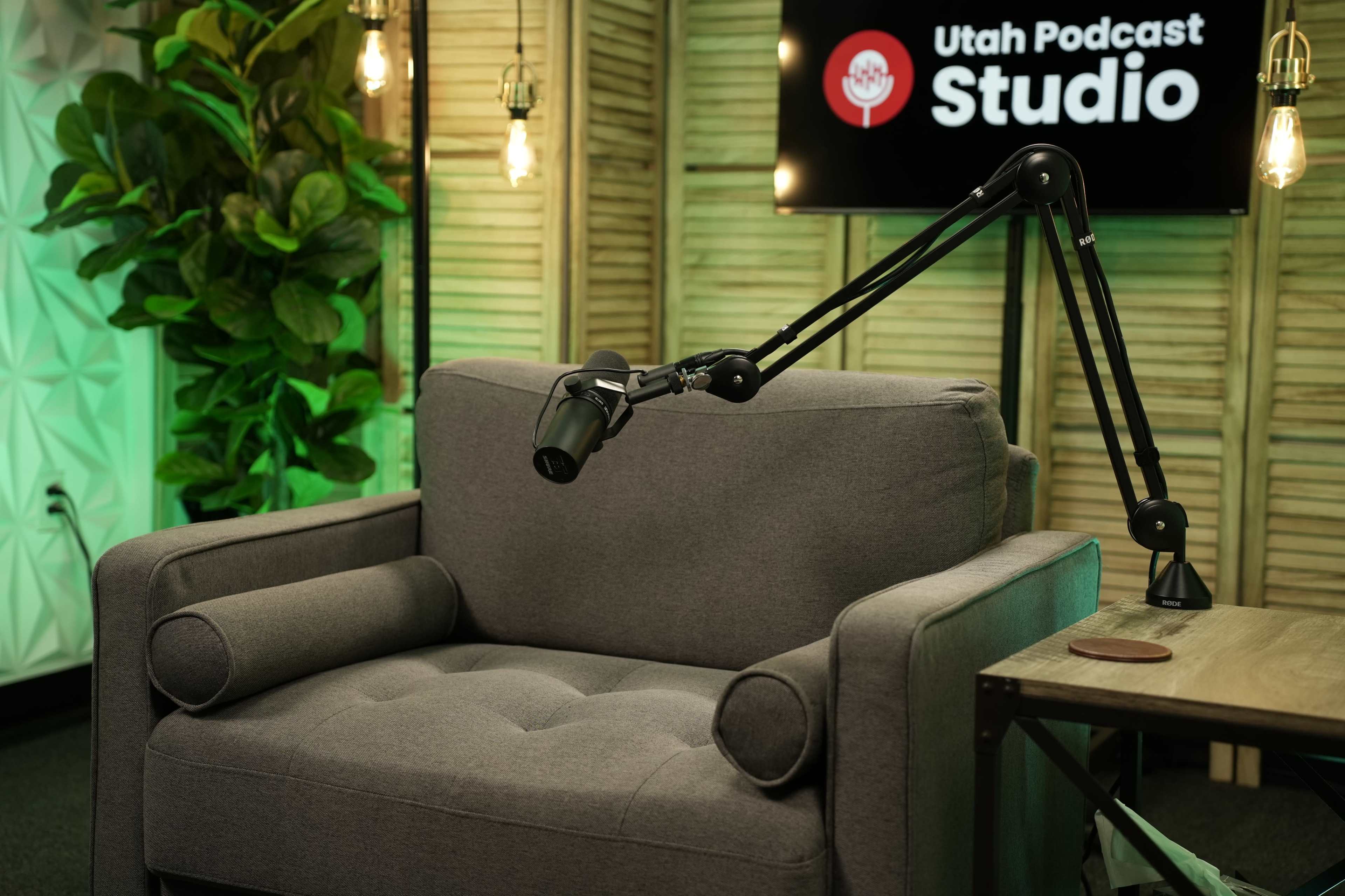 The image shows a grey armchair with a microphone attached to an adjustable arm, set in a podcast studio featuring a wooden backdrop and hanging lights.
