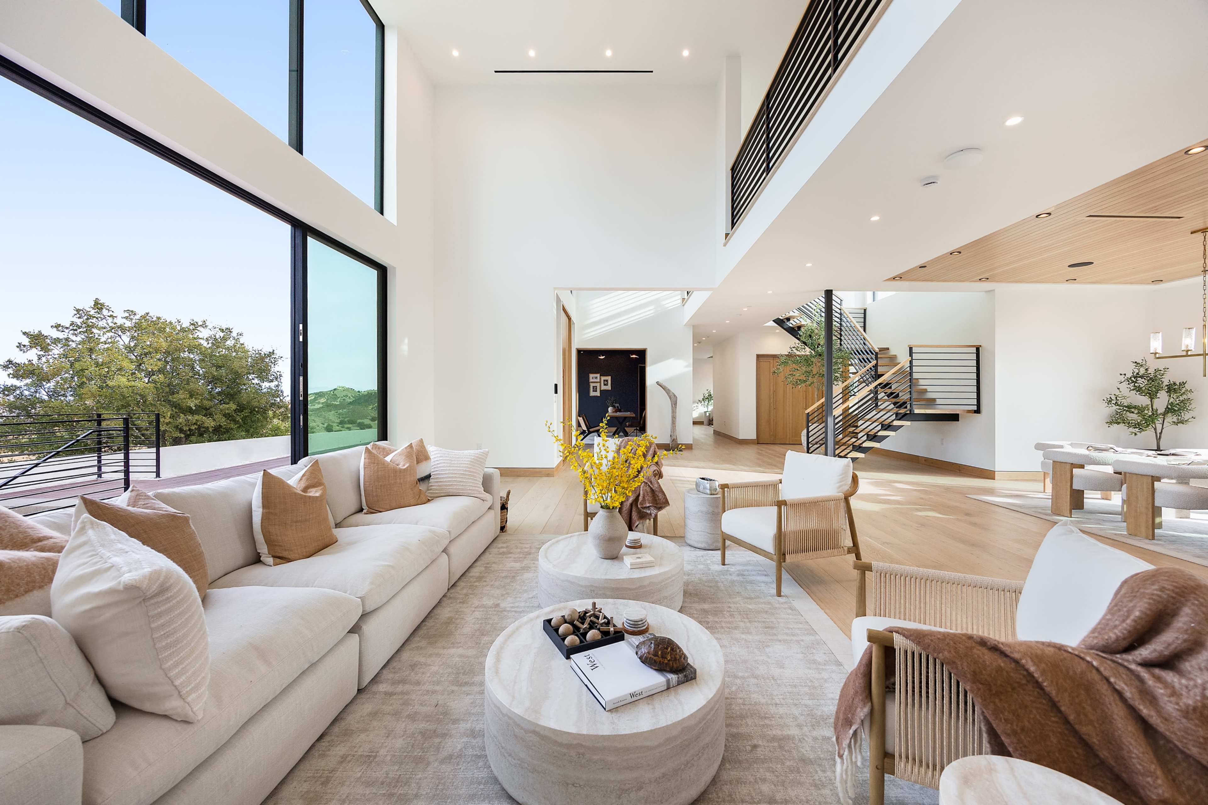A modern living room features a large sofa, light-colored decor, and floor-to-ceiling windows that showcase a view of greenery outside.