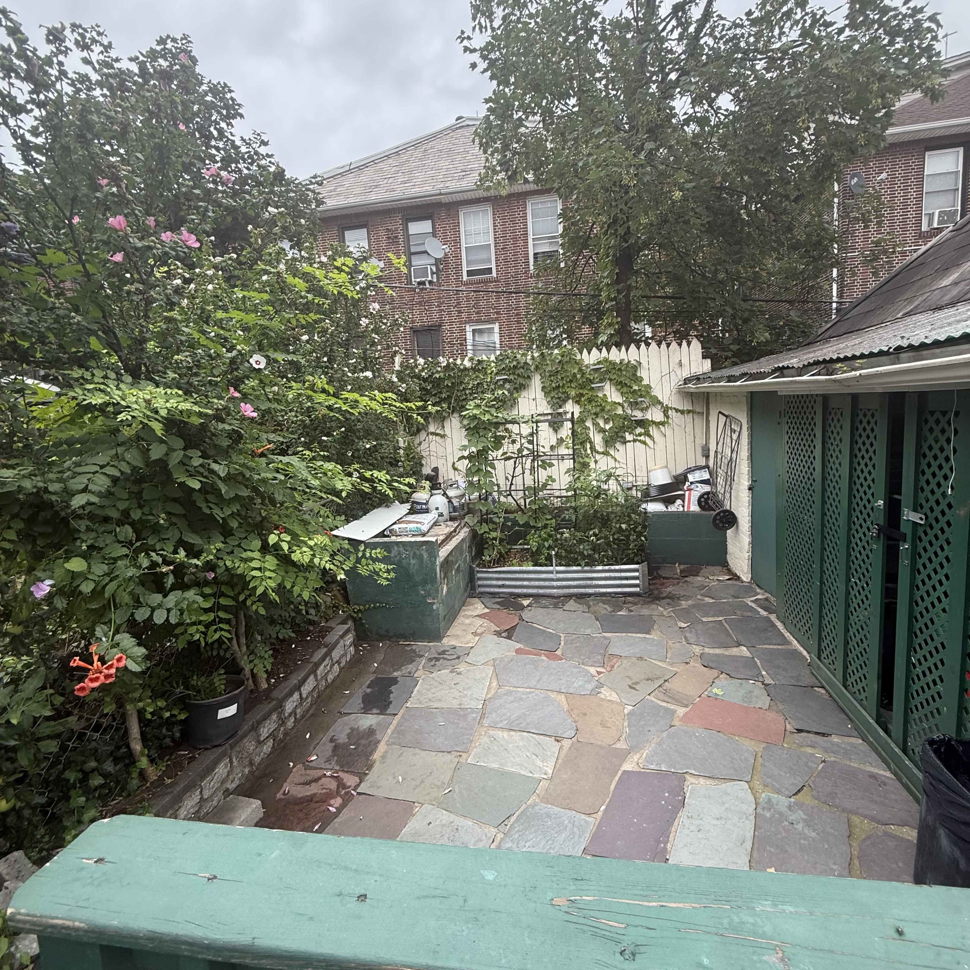 The image shows a small backyard area with stone paving, lush greenery, and a fence behind a residential building.