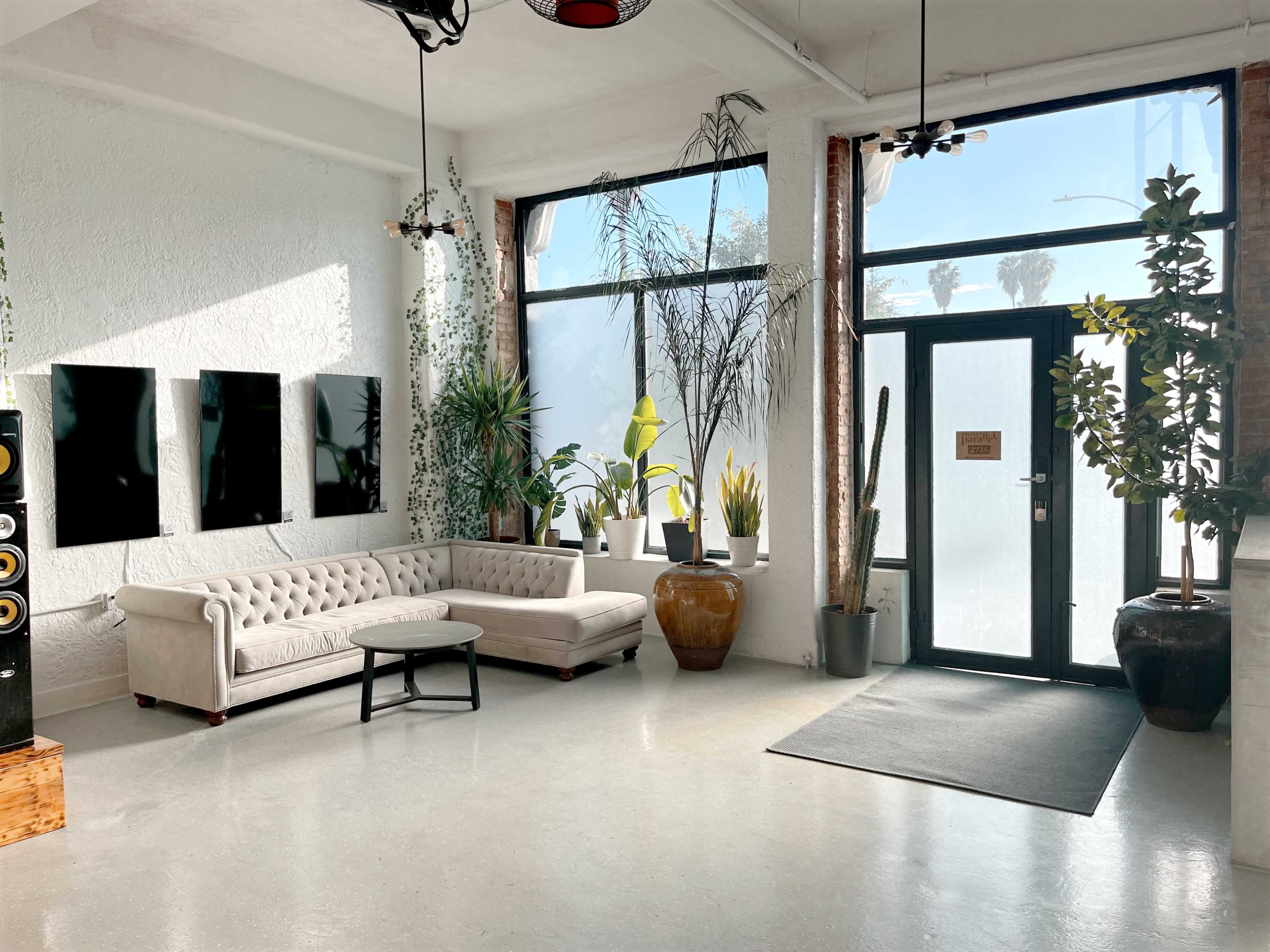 A bright, modern interior with a light-colored sofa, a round table, and large windows surrounded by various plants.
