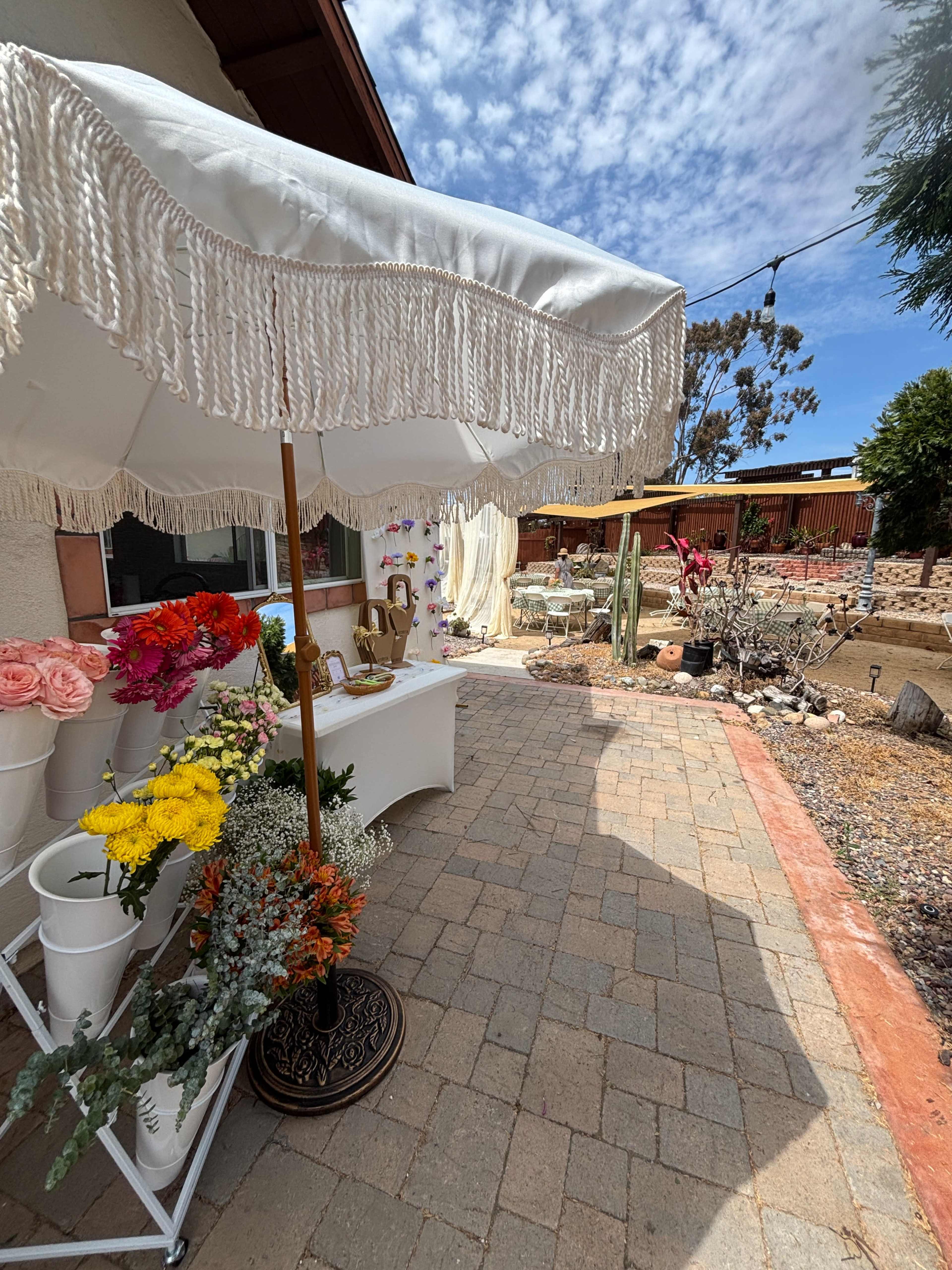 A white parasol with fringe stands near a pathway lined with colorful flowers and leads to a decorated outdoor space.