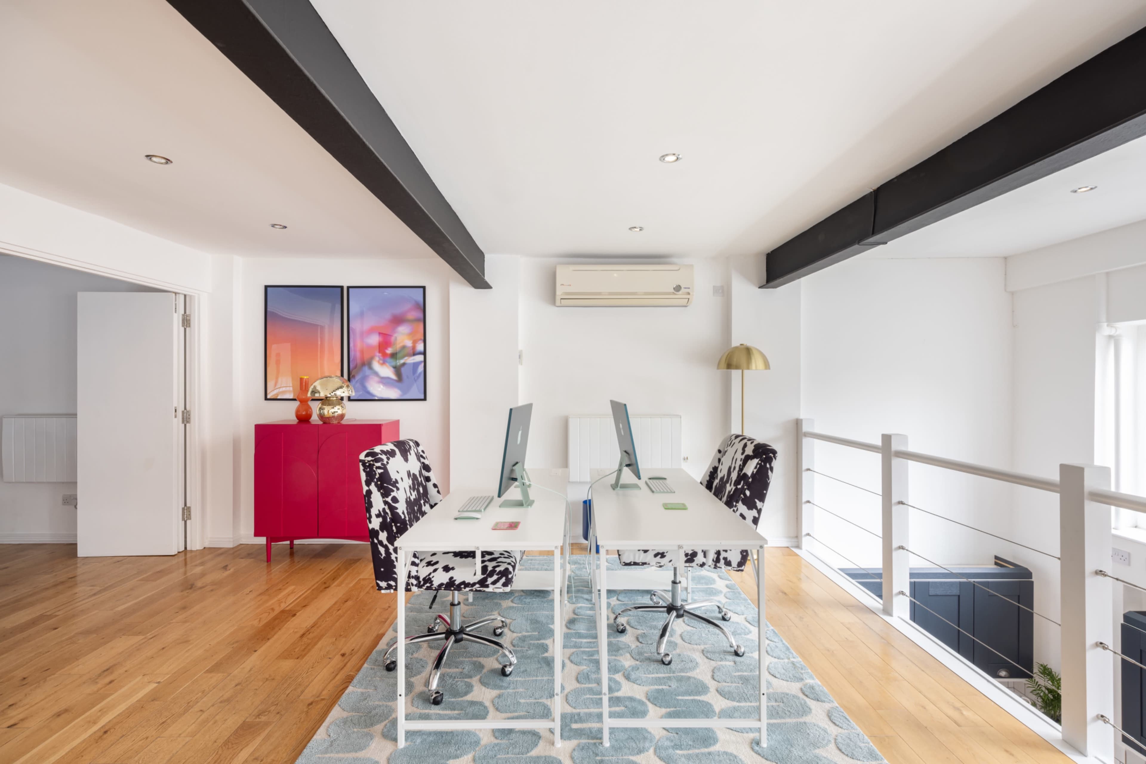 The image shows a minimalist office space with two desks facing each other, a colorful chest of drawers, and large windows letting in natural light.
