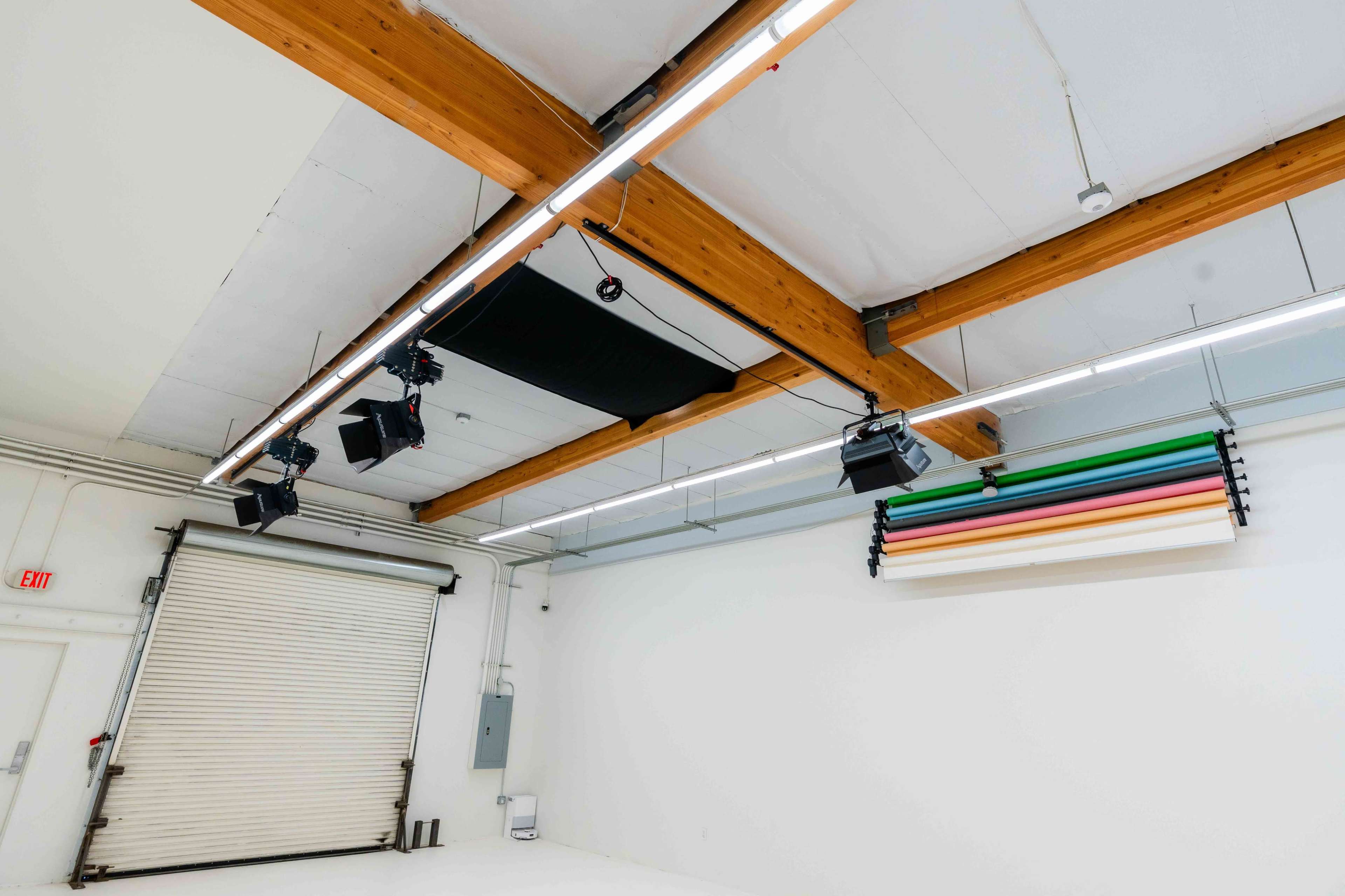 The image shows an empty studio space with wooden beams, overhead lighting, and a roll-up door, featuring a color backdrop system on one wall.