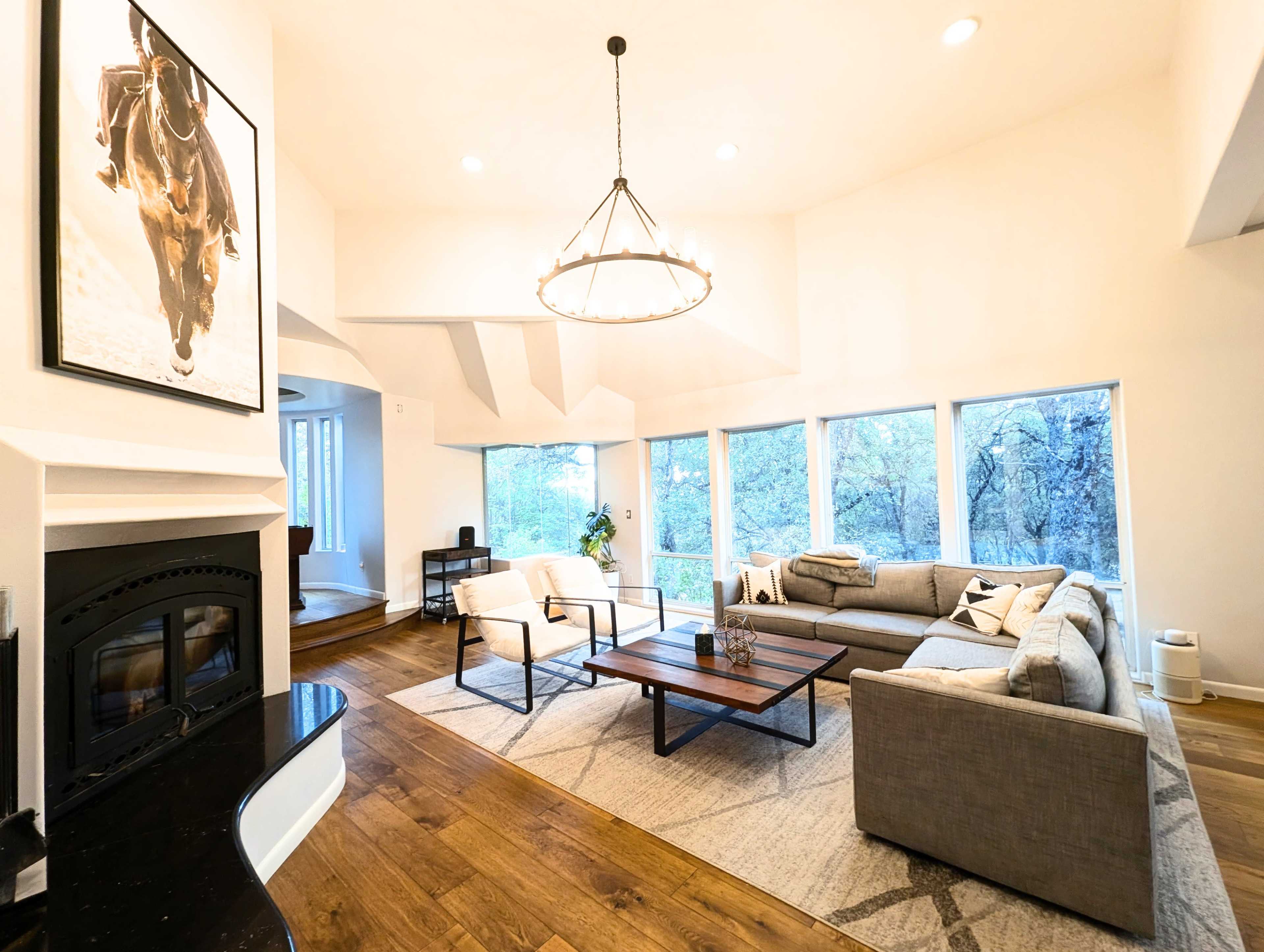 A spacious living room features large windows, a sectional sofa, and a central coffee table under a circular chandelier.