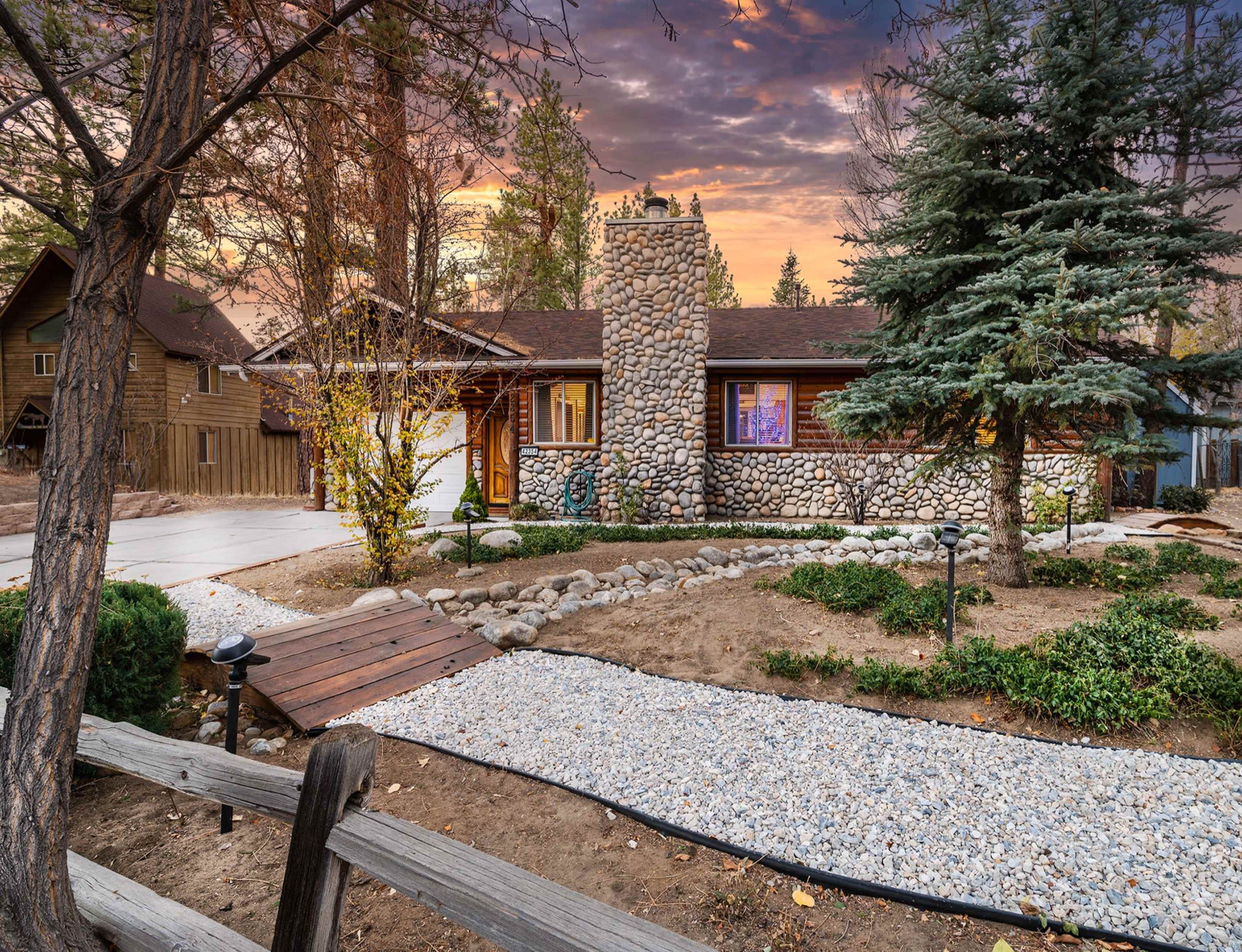A ranch-style house with a stone fireplace and landscaped yard is set against a colorful sunset sky.