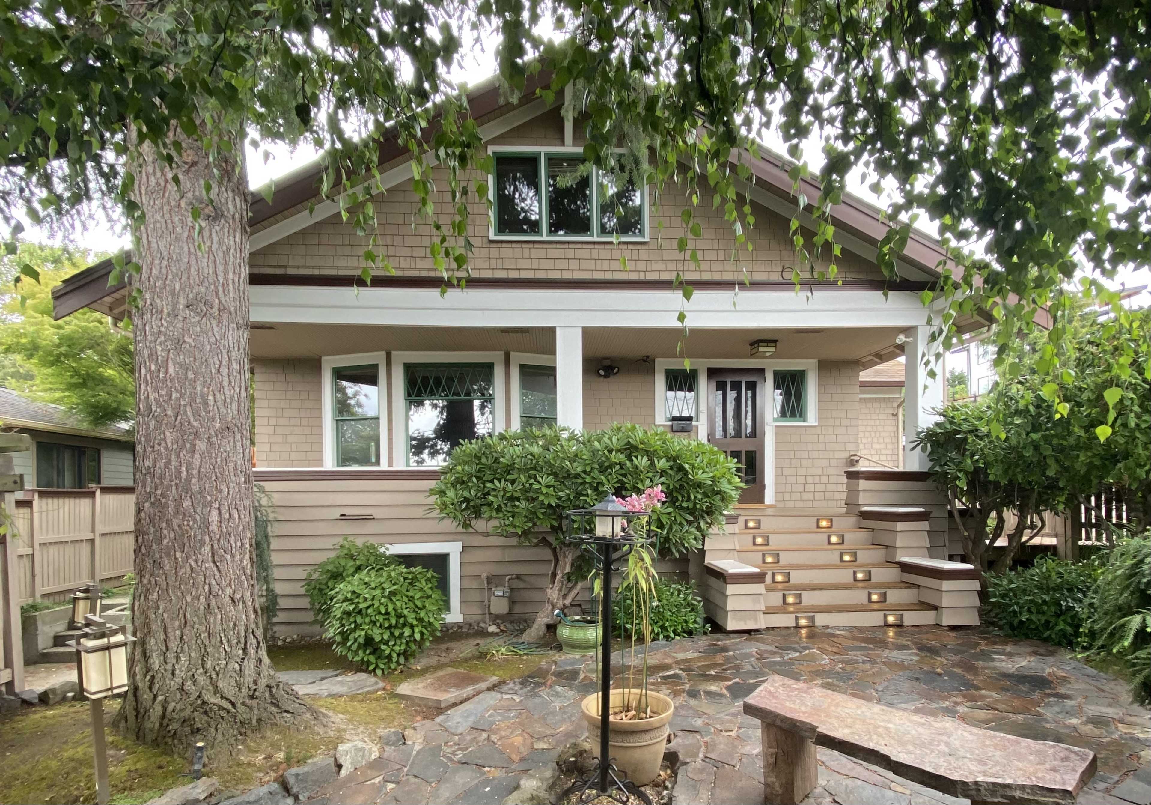 A two-story house with a front porch, surrounded by lush greenery and stone pathways.