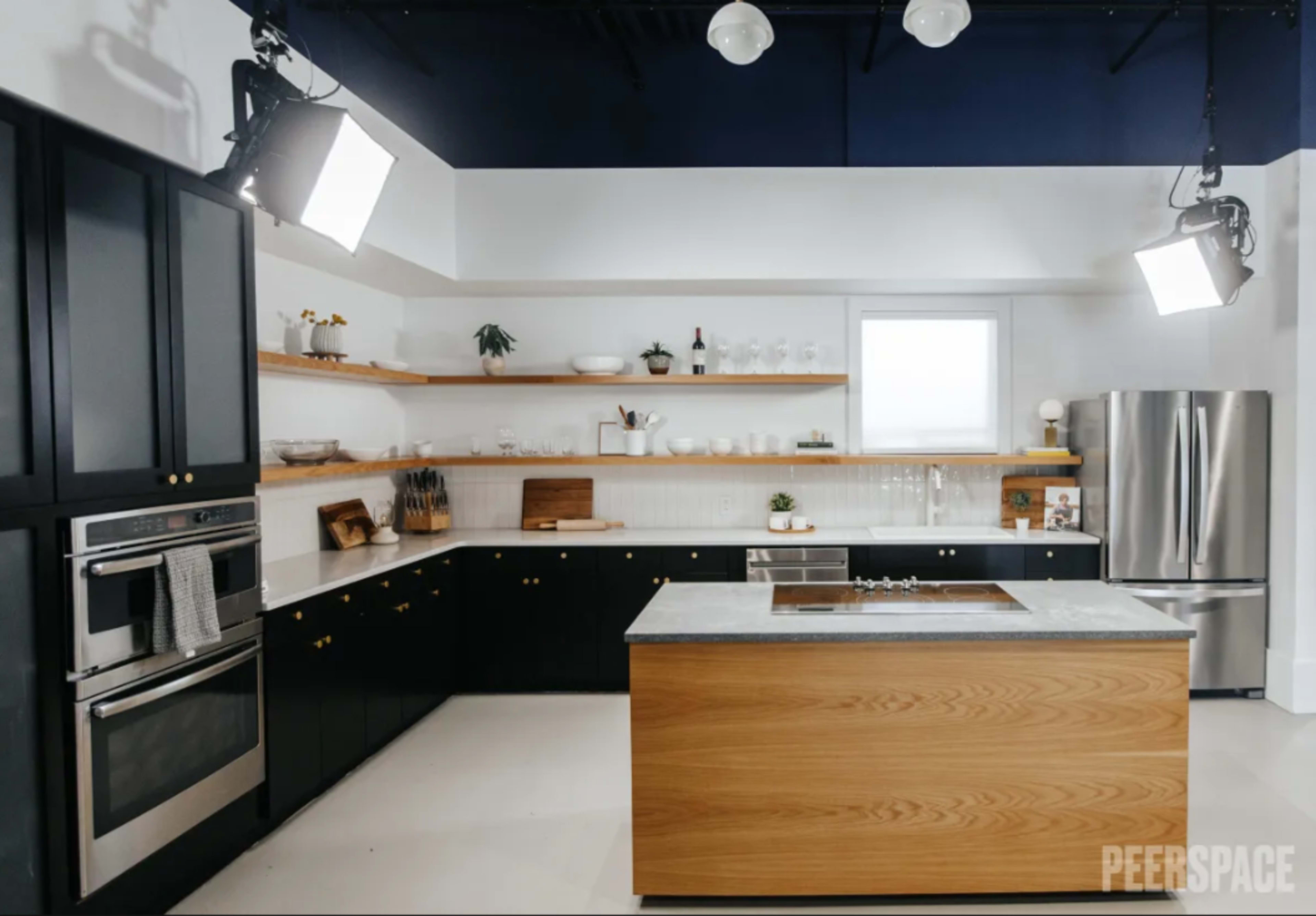 A modern kitchen featuring black cabinetry, a central wooden island, and stainless steel appliances, with shelves displaying dishware.