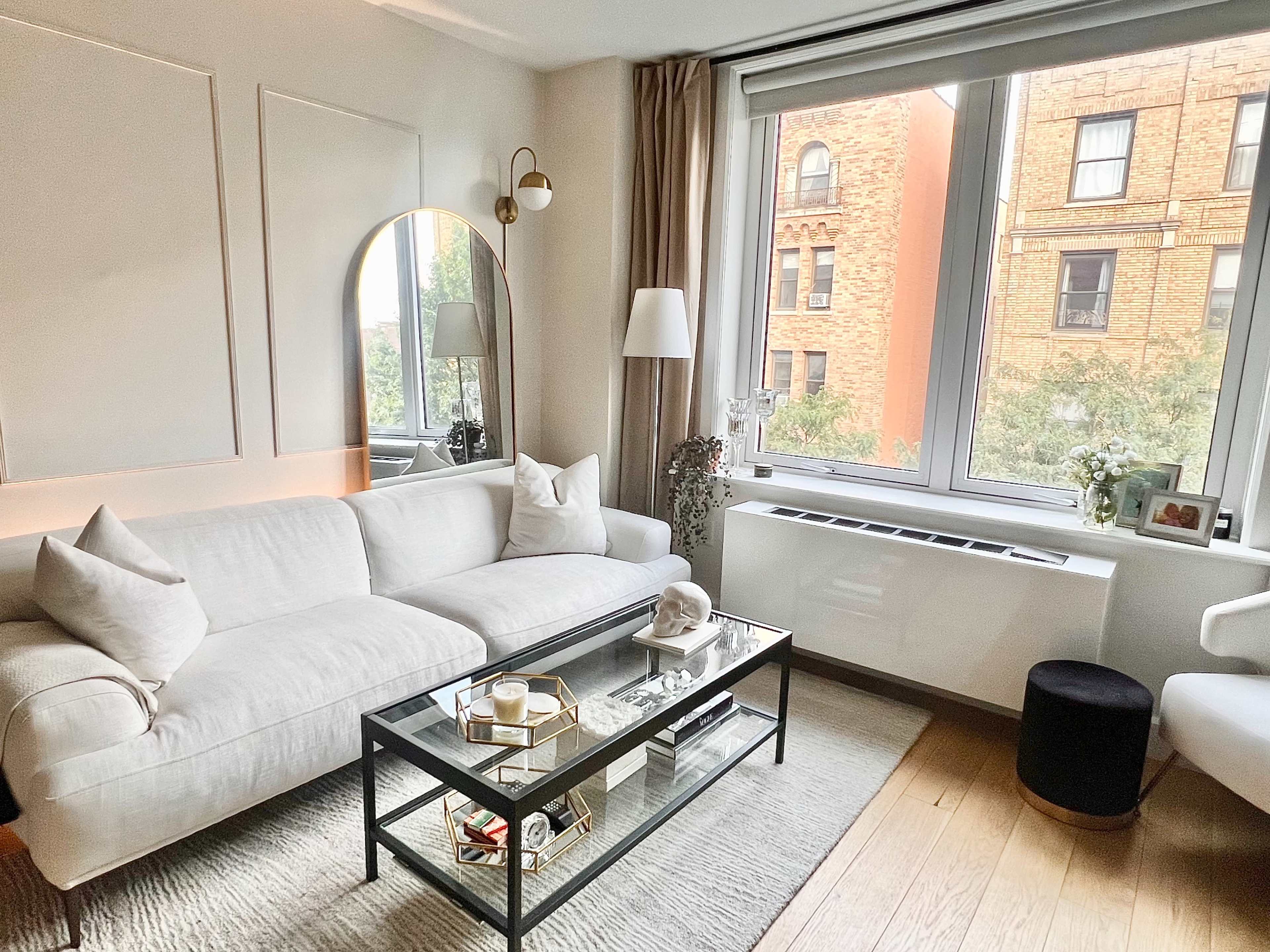 A neatly arranged living room featuring a white sofa, a glass coffee table, and large windows overlooking a brick building.