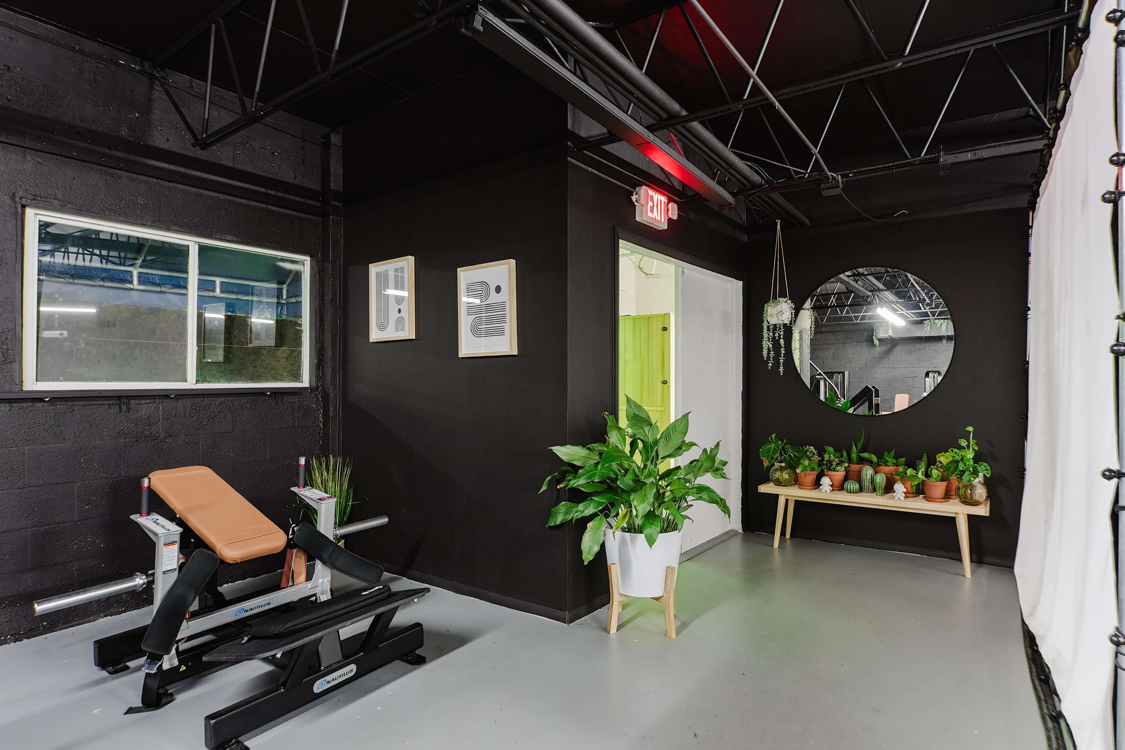 The image shows a modern fitness studio featuring a weight bench, various potted plants, and a round mirror on a black wall.