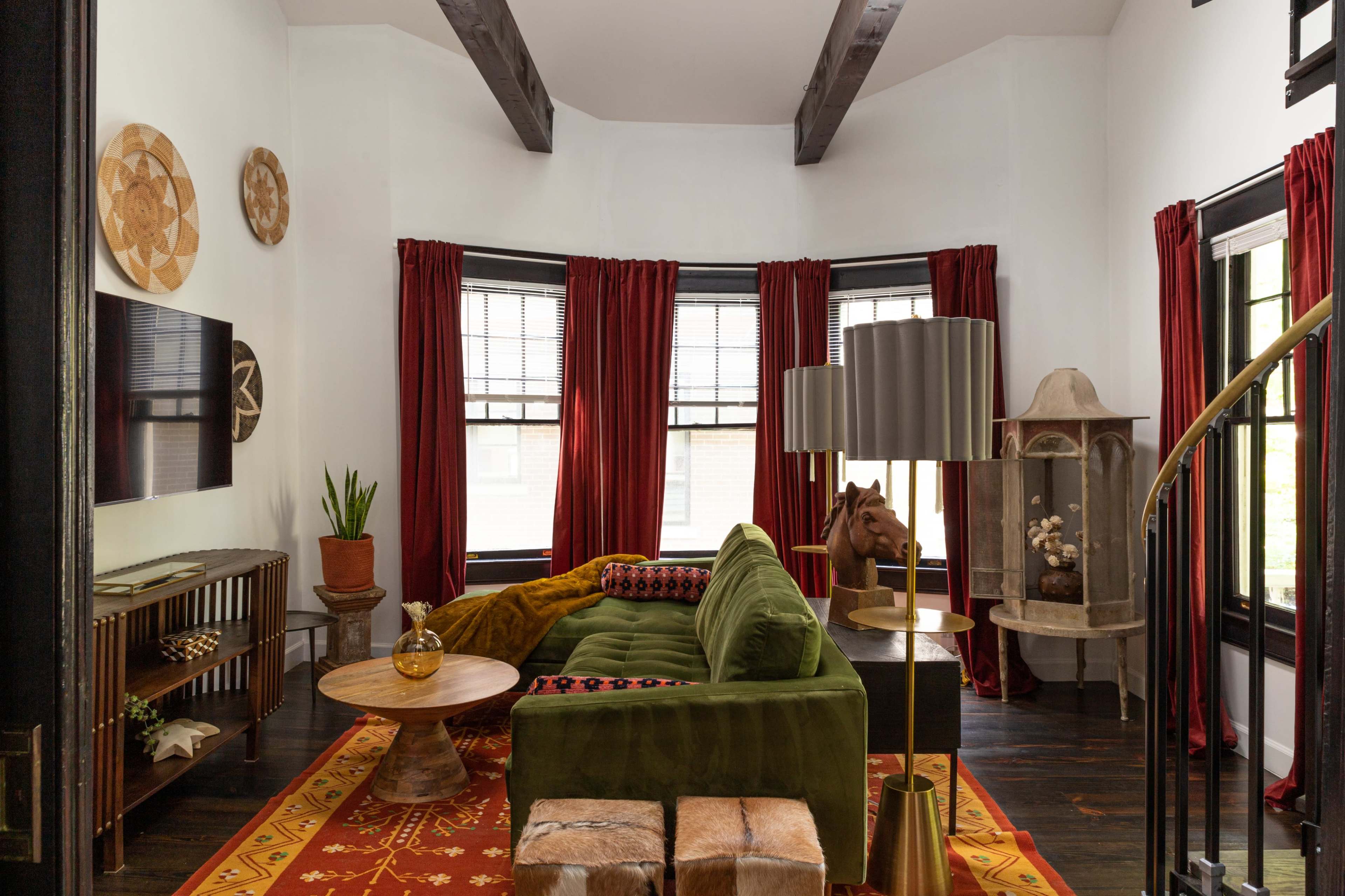 A living room with a green sofa, a round coffee table, and large windows draped with red curtains, featuring wooden beams on the ceiling.