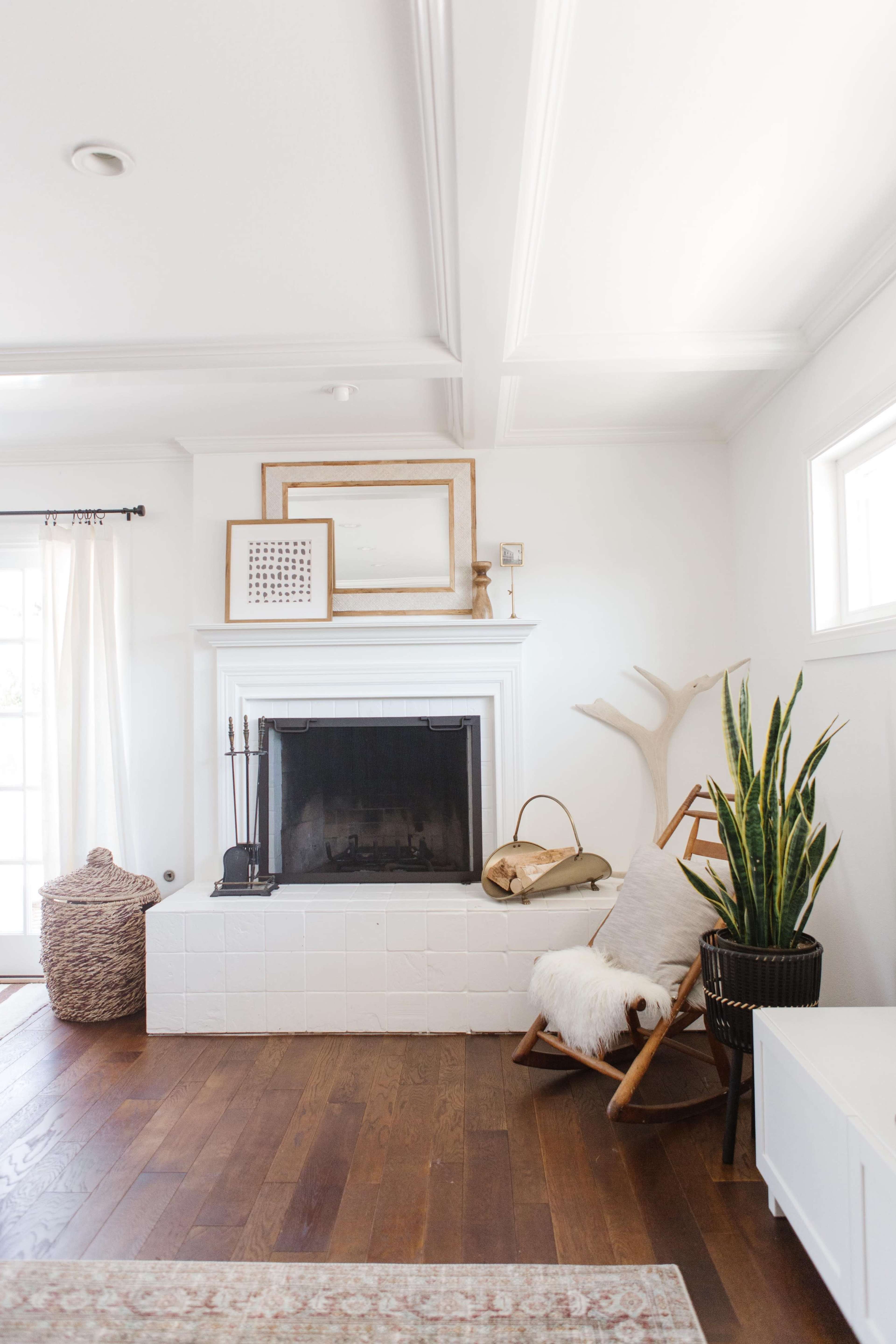 A modern living room with a white tiled fireplace, a wooden floor, and a decorative plant beside a chair and a wicker basket.