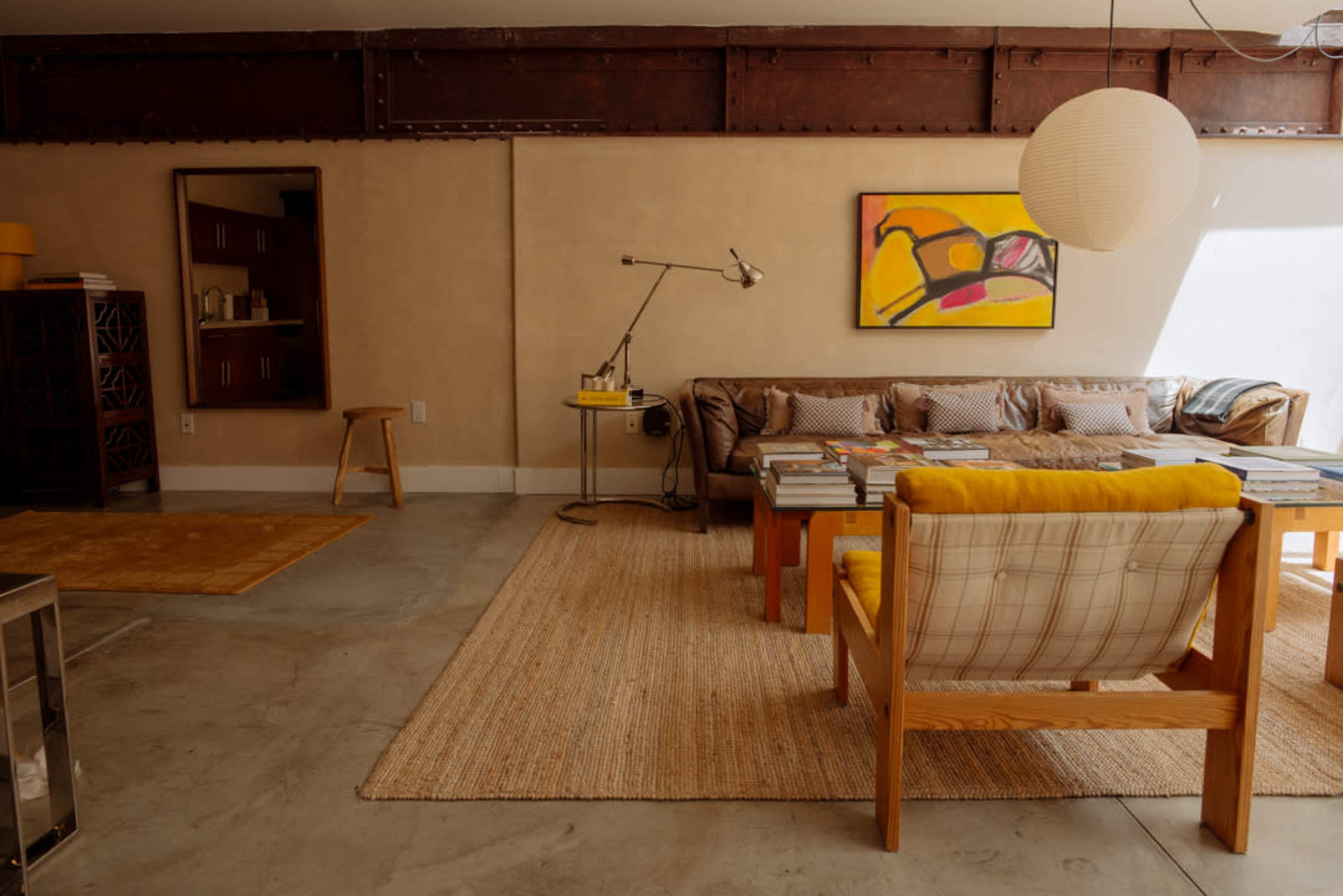 The image shows a modern living room featuring a brown leather sofa, a coffee table stacked with books, a yellow accent chair, and an abstract painting on the wall.