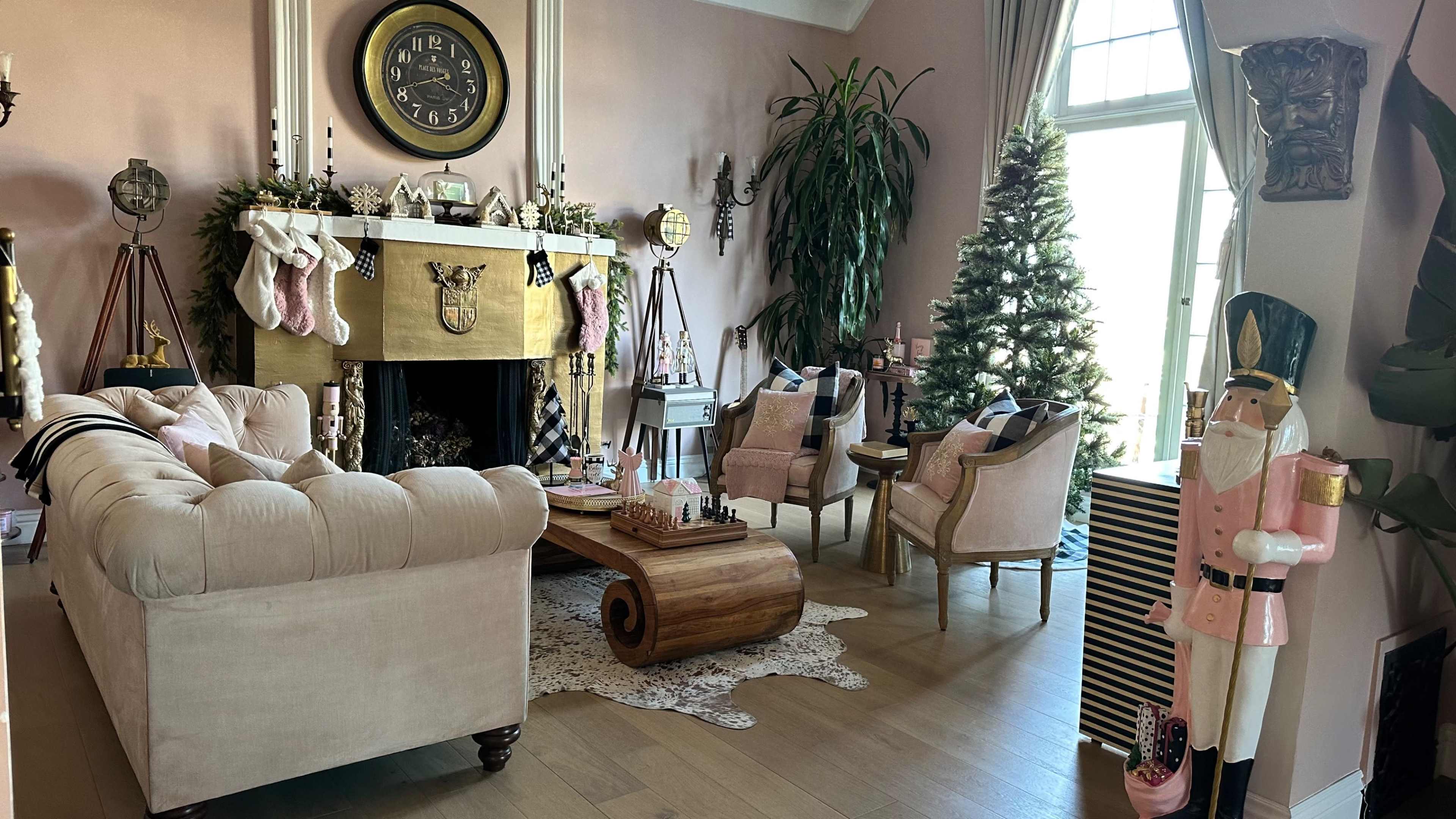 A cozy living room features a beige couch, a decorative fireplace adorned with stockings, a Christmas tree, and two armchairs, along with festive decor such as a nutcracker and holiday ornaments.