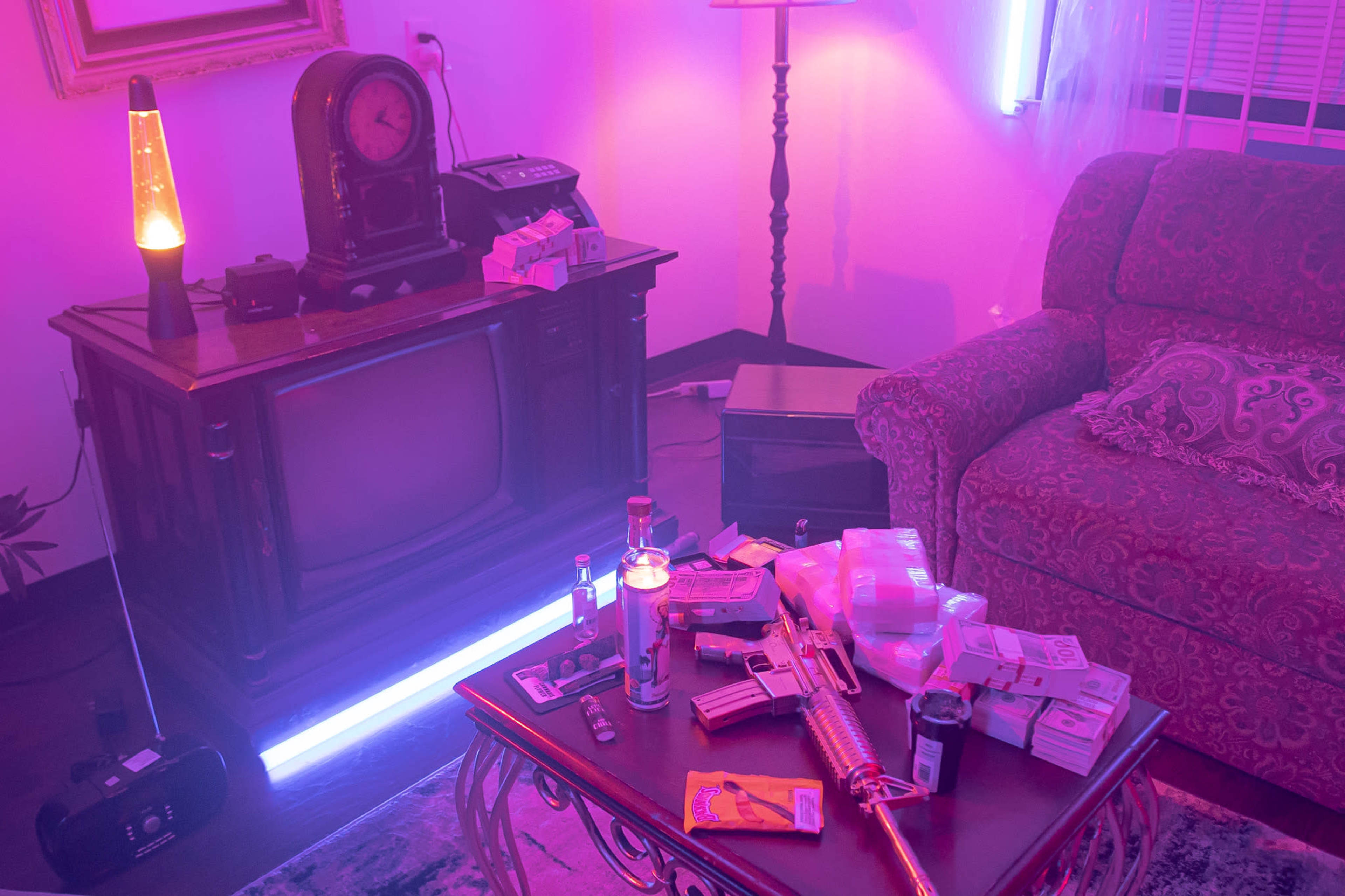 A cluttered living room features an old television set, a vintage clock, a lava lamp, and a coffee table covered with various items, including an airsoft gun and packs of cigarettes.