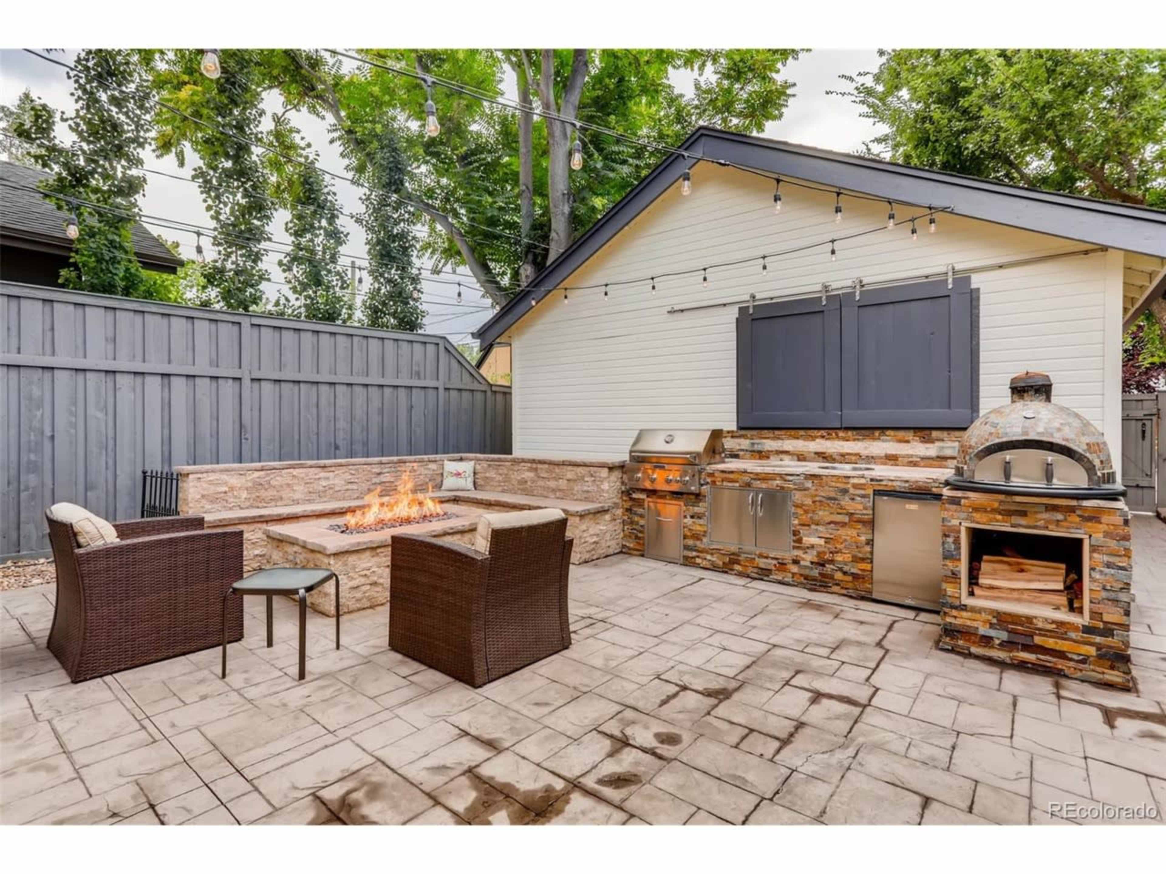 The image shows an outdoor patio area with a stone fire pit, seating arrangement, a grill, and a pizza oven against a backdrop of a gray fence and trees.