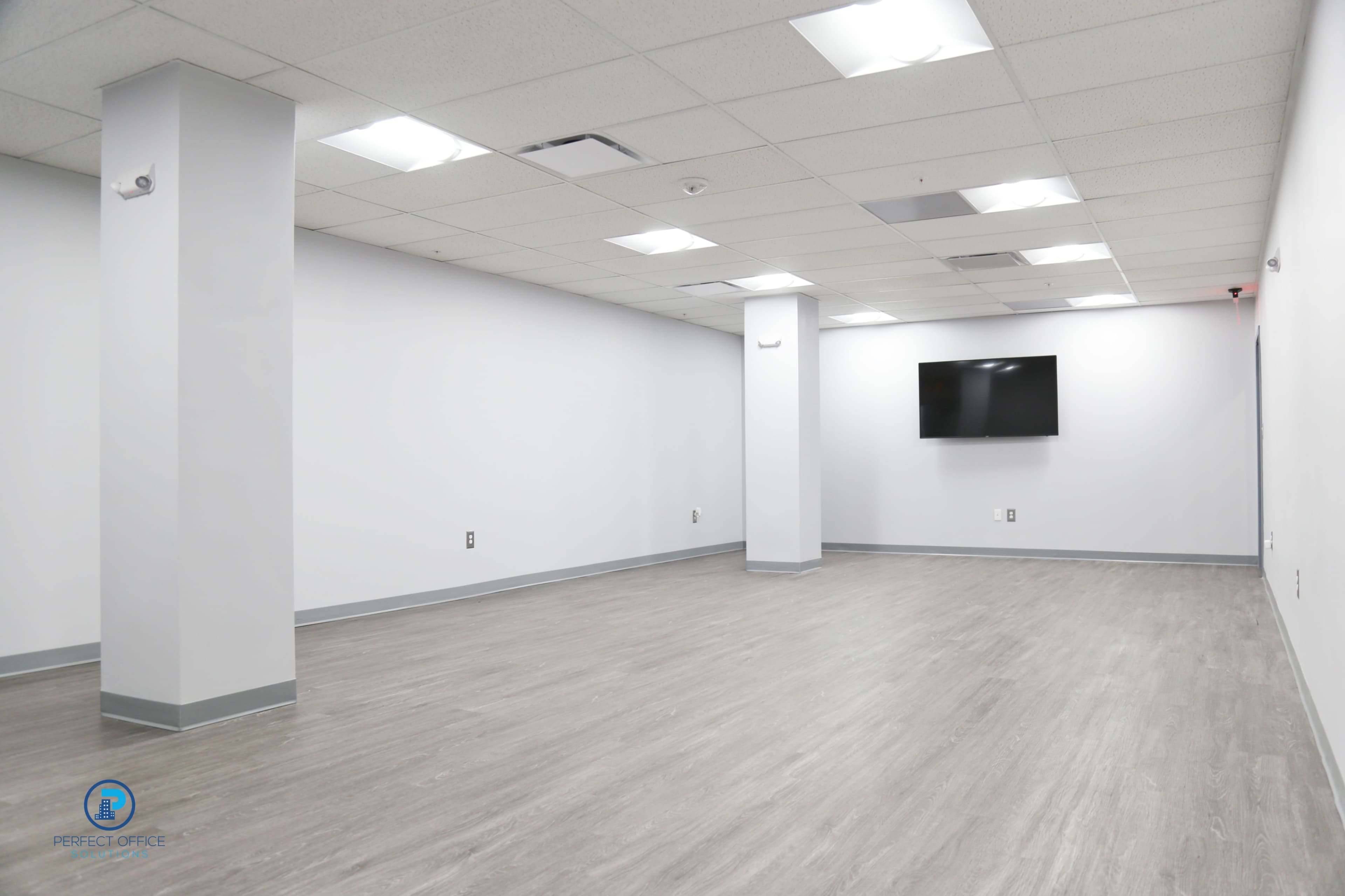An empty, well-lit room with gray flooring, white walls, a wall-mounted television, and no furniture.
