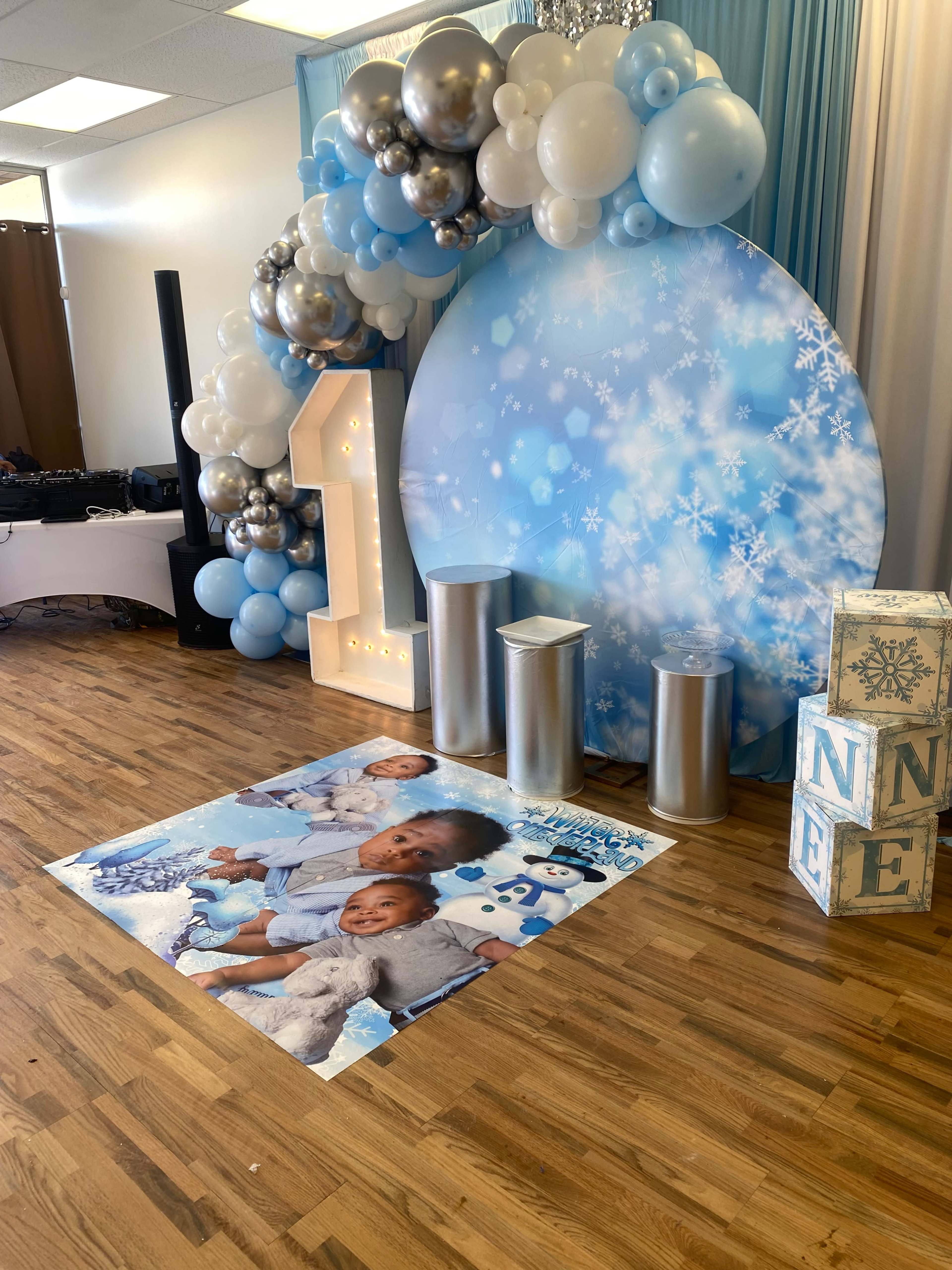 A festive decoration area for a one-year-old's birthday celebration, featuring a large number "1" illuminated with lights, an arch of blue and silver balloons, and a floor mat with photographs of children.