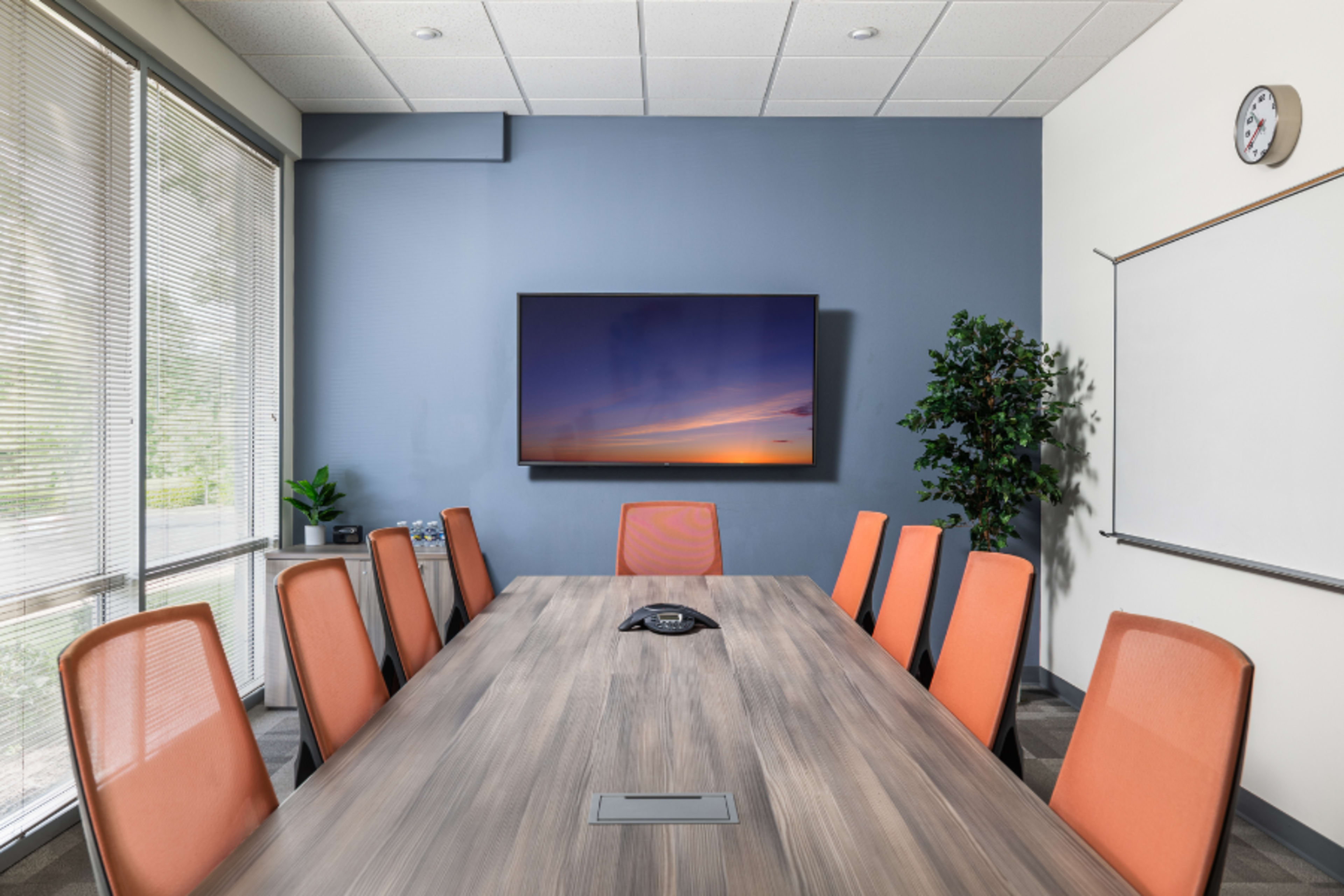 A modern conference room features a long wooden table surrounded by orange chairs, with a large screen mounted on the wall and a whiteboard nearby.