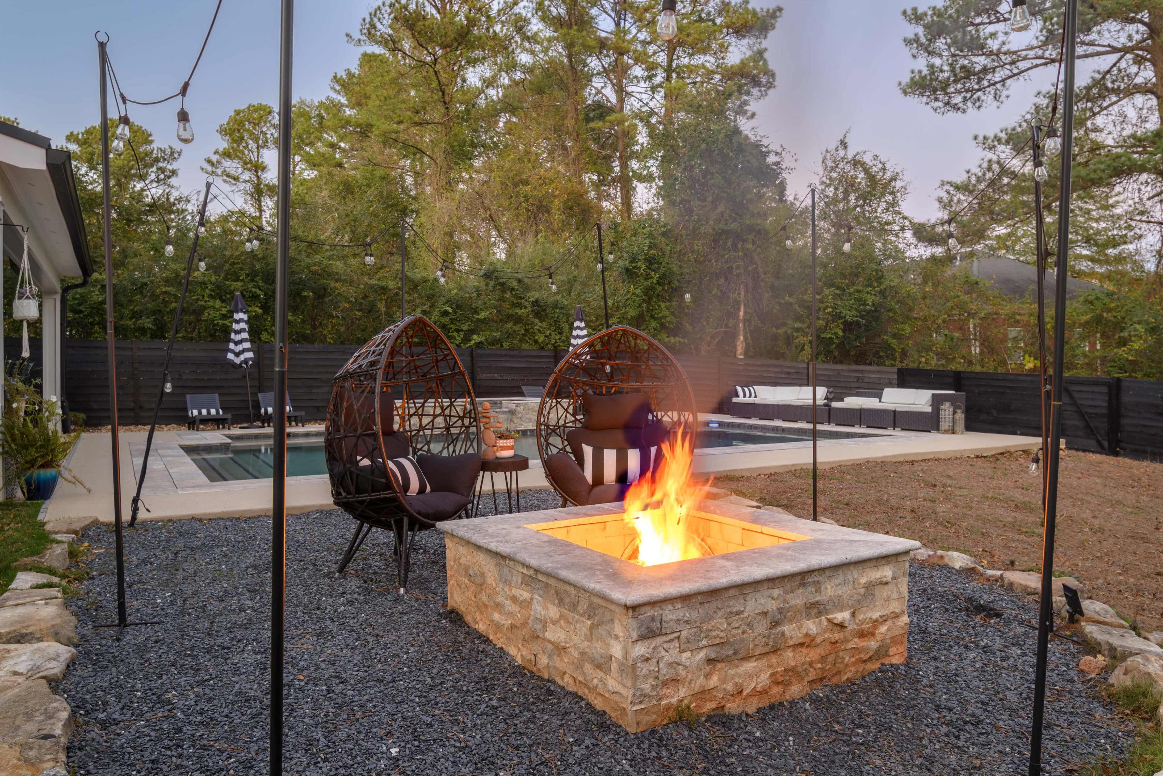 The image shows a backyard area with two hanging chairs near a stone fire pit, surrounded by landscaping and a swimming pool in the background.