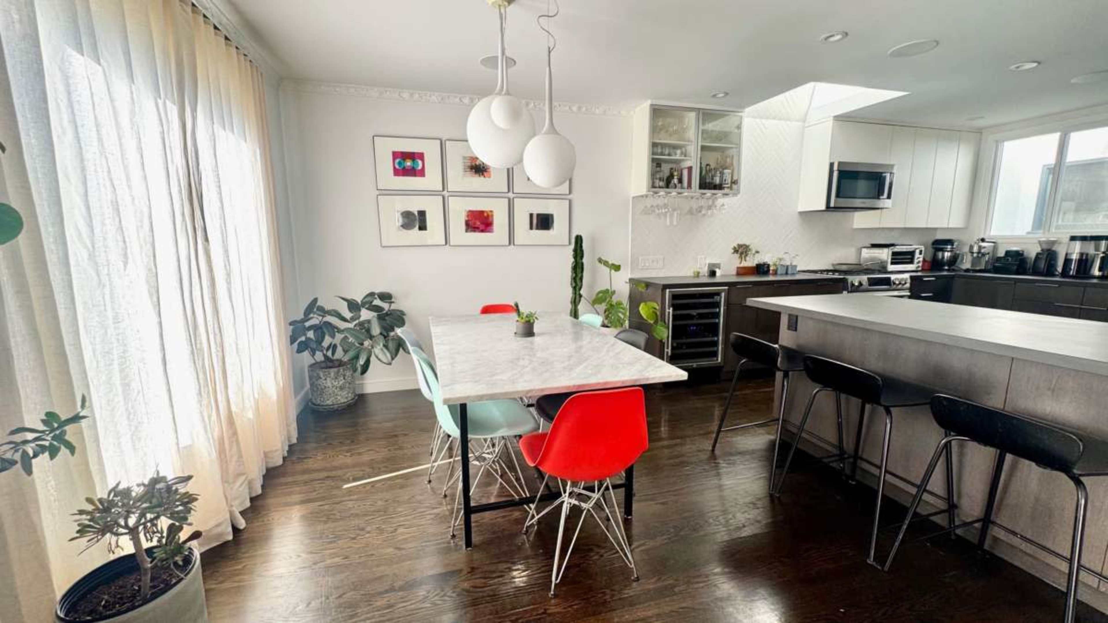 The image shows a modern kitchen and dining area featuring a marble-topped table with colorful chairs, potted plants, and large windows allowing natural light to enter.