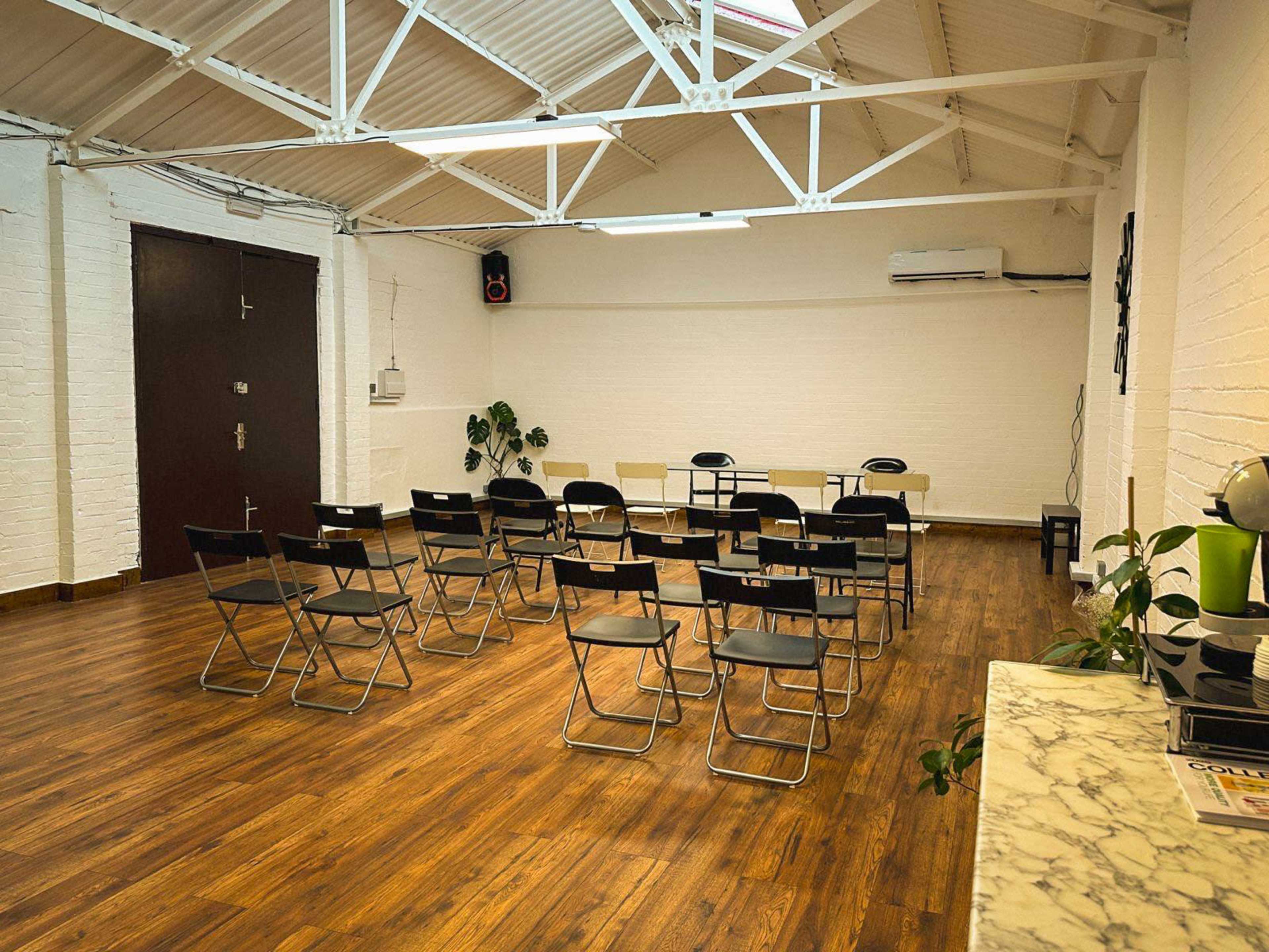The image shows a spacious room with wooden flooring, featuring several black folding chairs arranged in rows facing a table at the front.