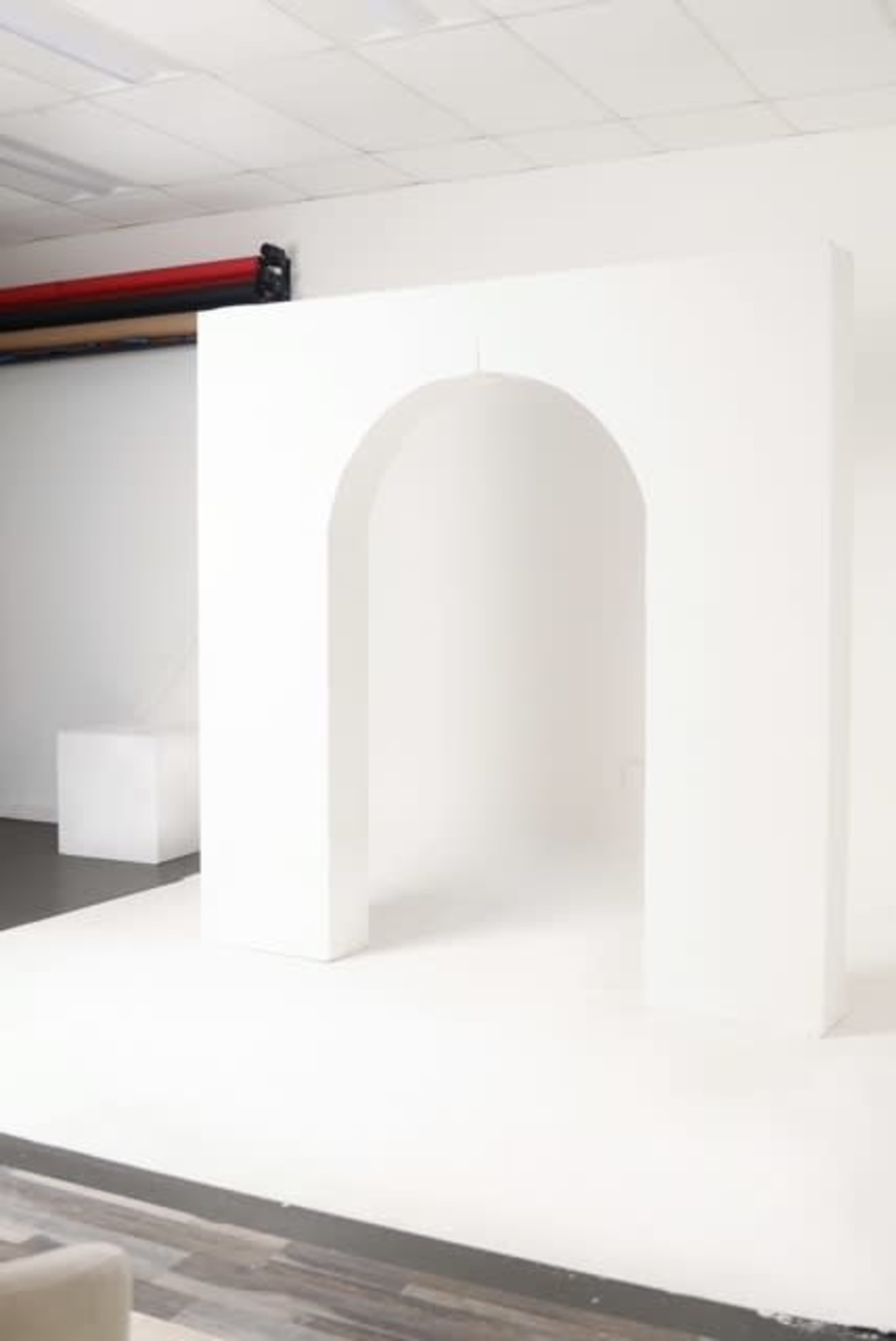 The image shows a minimalist white archway set against a plain white background in a studio space.