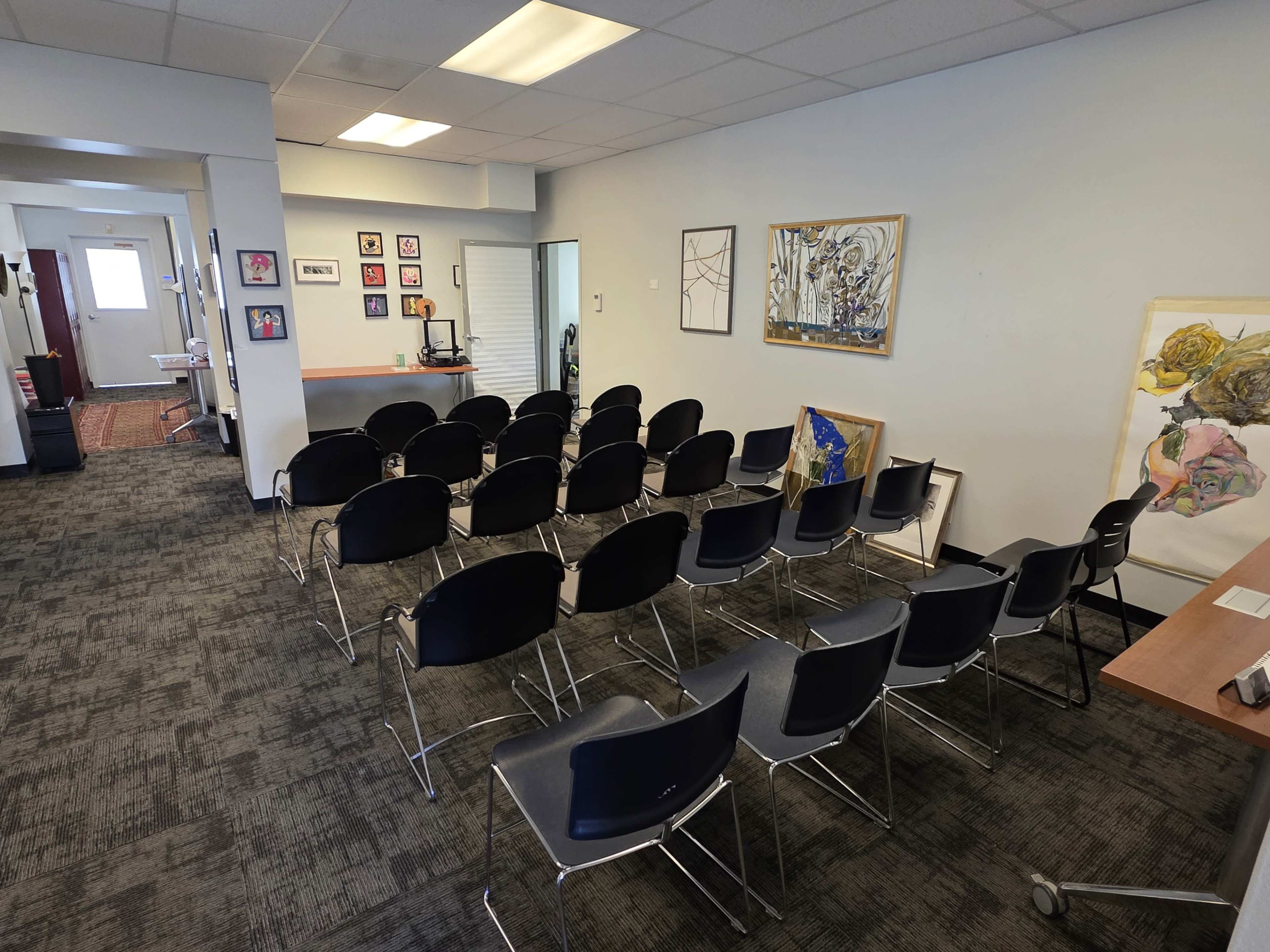 The image shows a small meeting room with several rows of black chairs arranged facing a wall adorned with framed artwork.