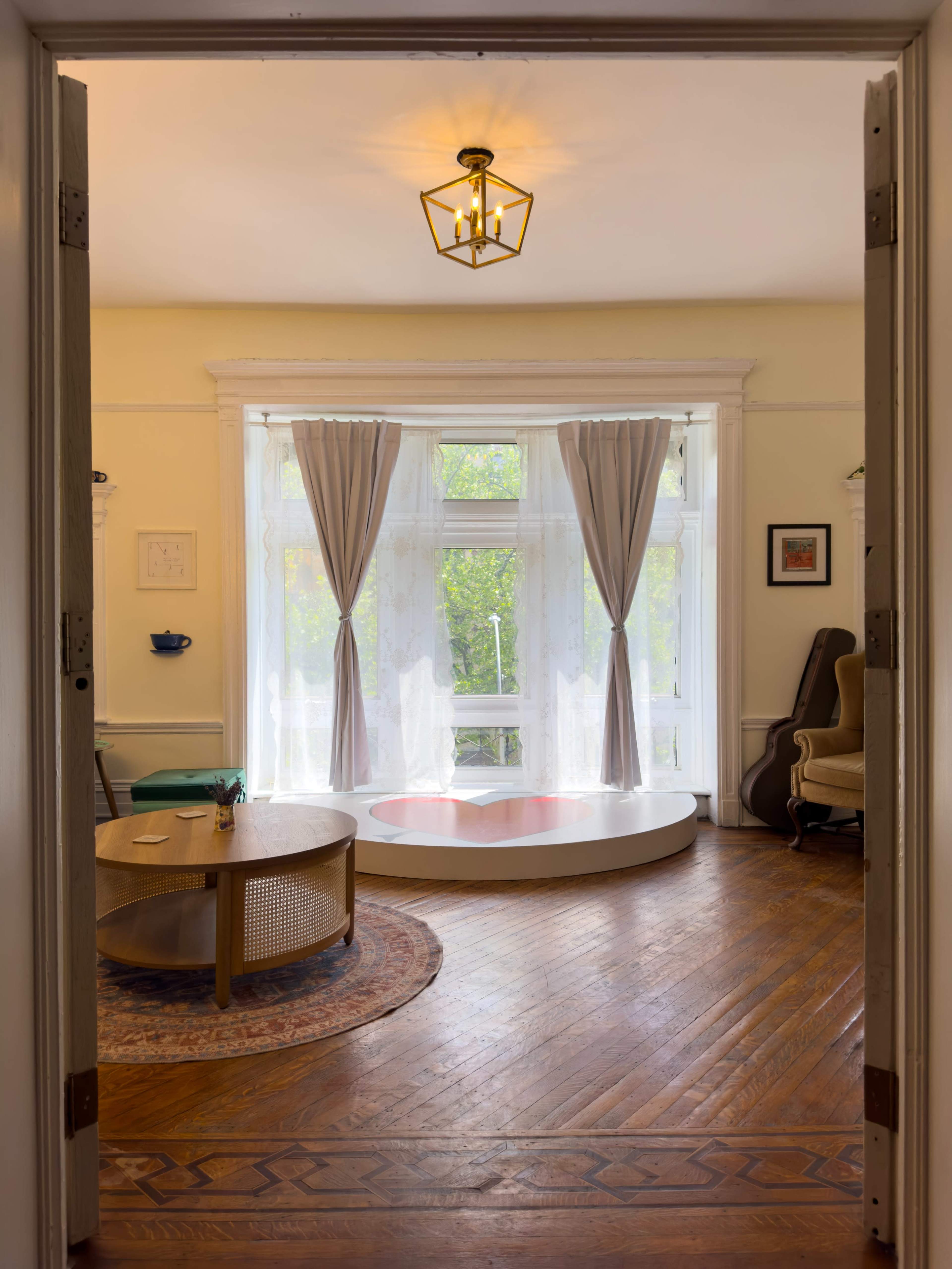 A bright, airy room features a round table and a circular, raised platform with a padded edge, framed by large windows dressed in sheer curtains.