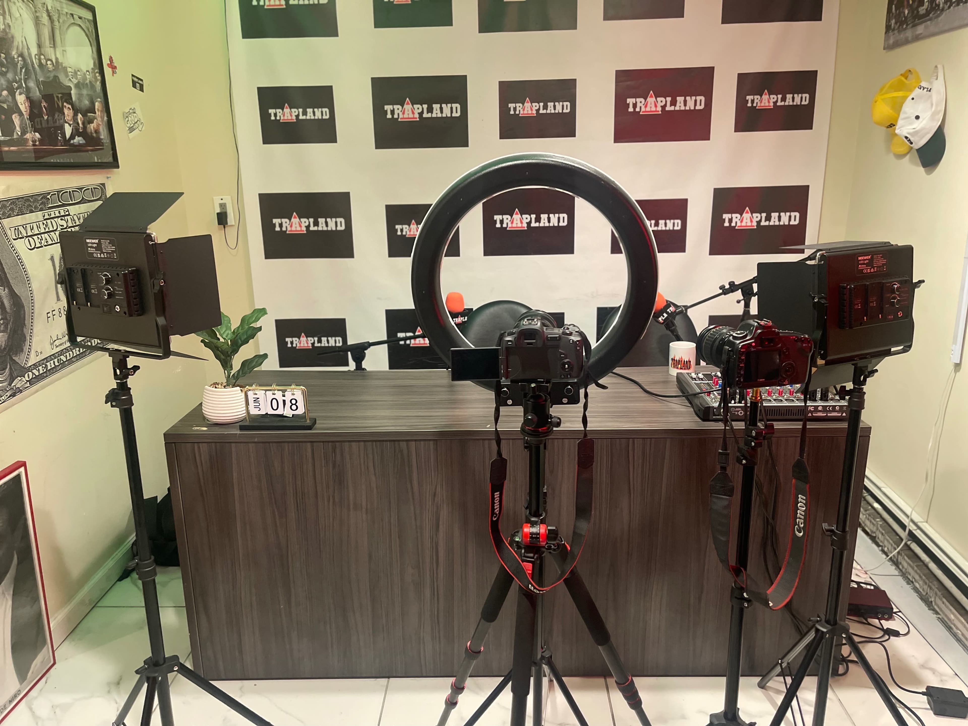 A video production setup featuring two cameras on tripods, a ring light, and a desk with microphones in a room decorated with branding elements.
