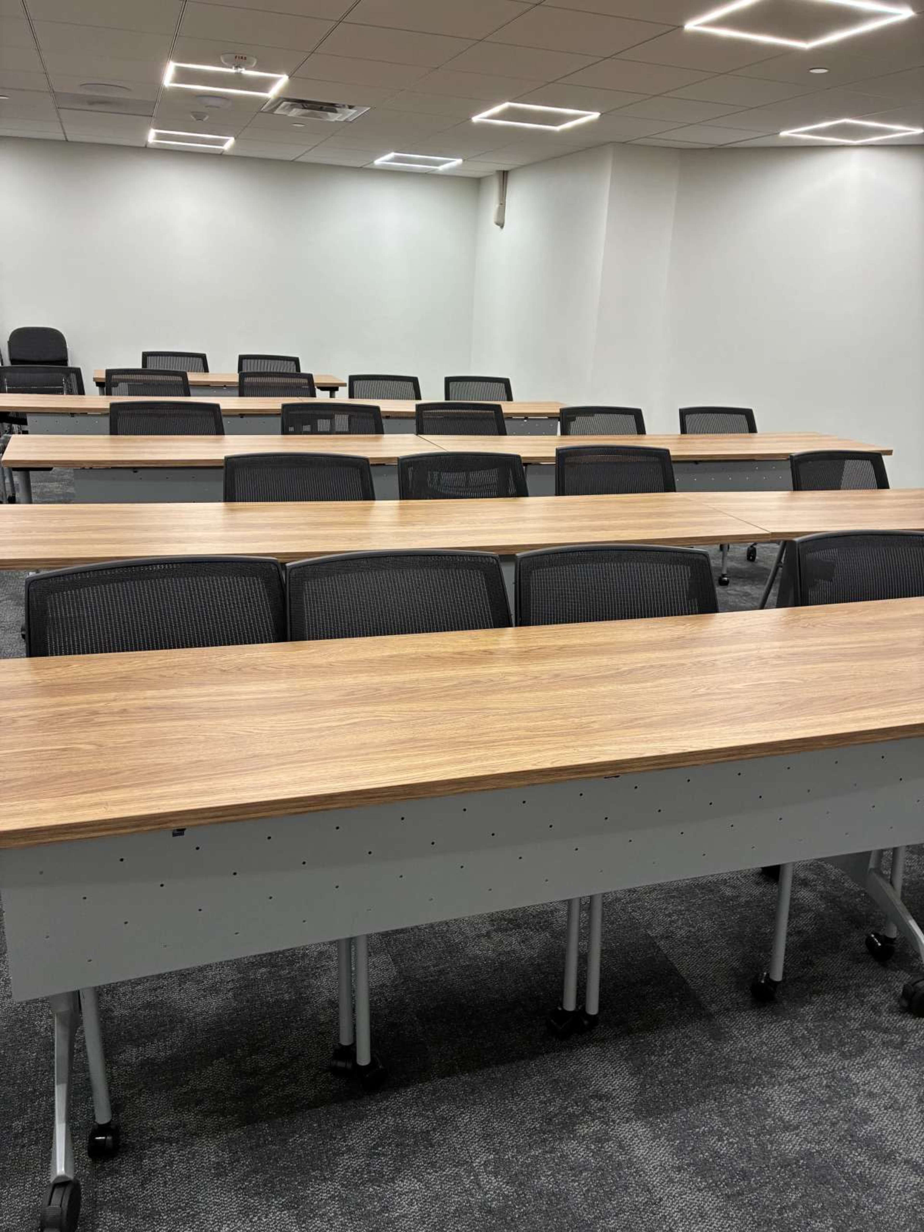 The image shows a classroom with rows of wooden tables and black rolling chairs arranged neatly.