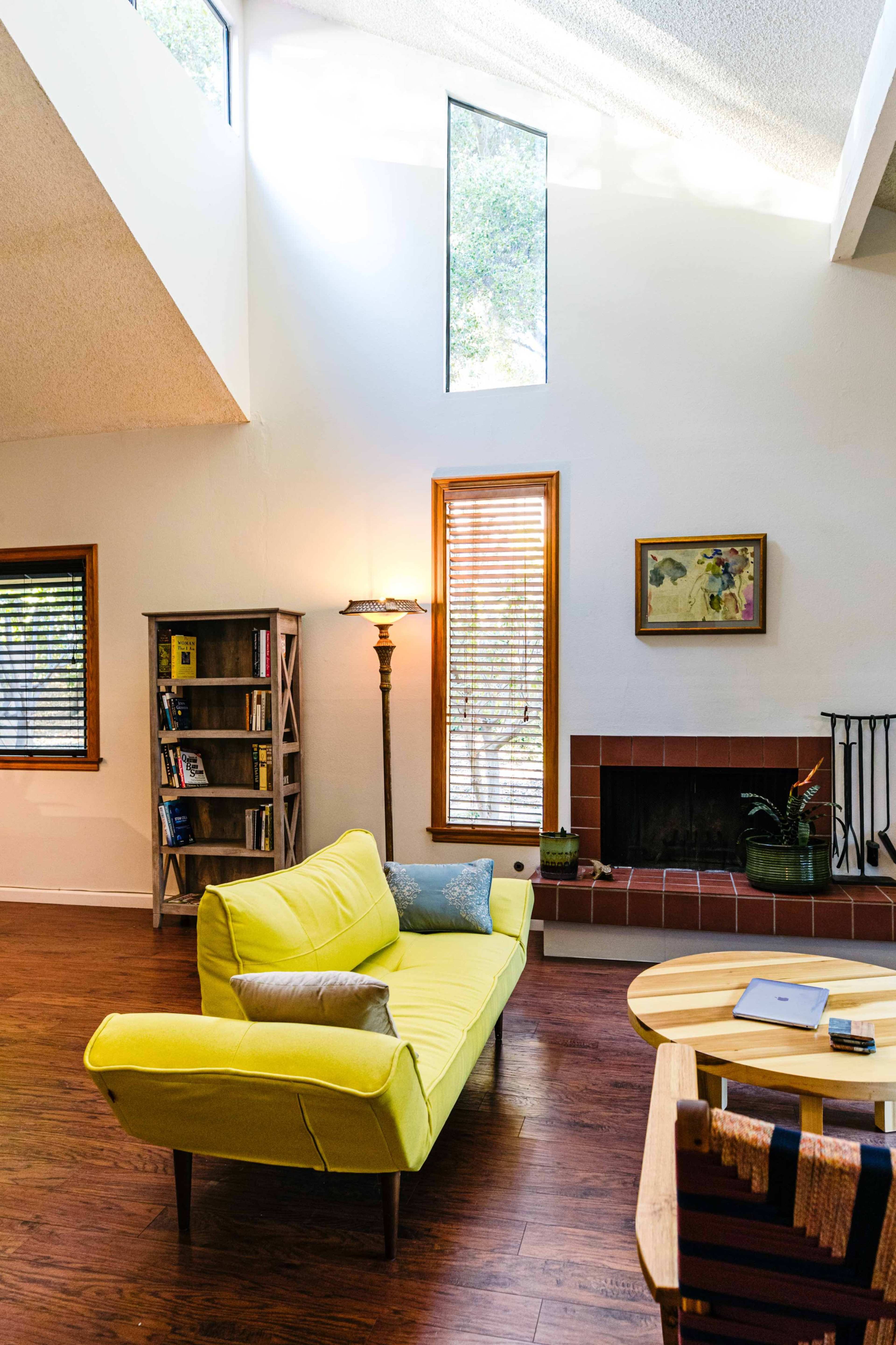 A bright living room features a yellow sofa, a wooden coffee table, a bookshelf, and a fireplace with natural light streaming through a skylight.