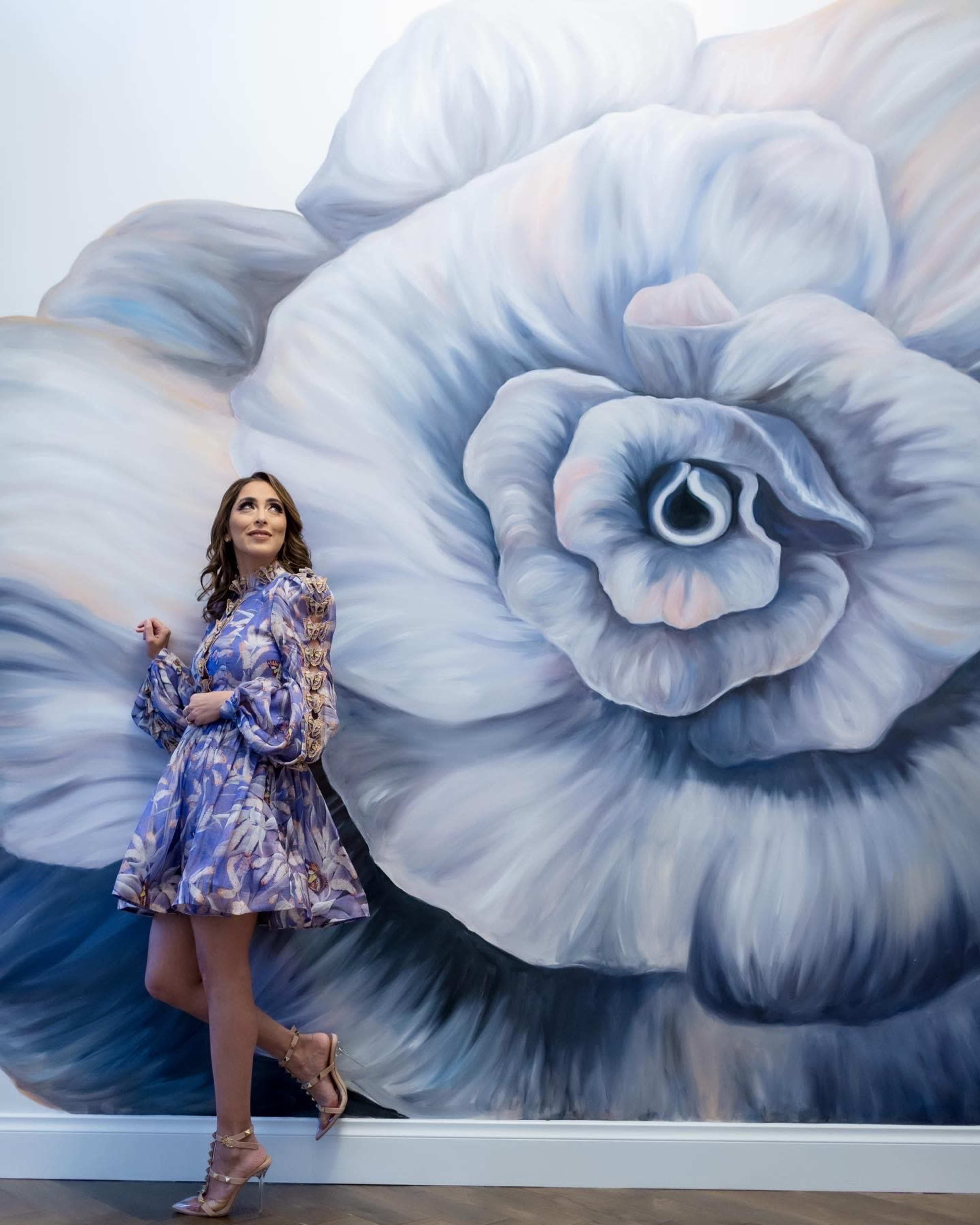 A woman poses beside a large mural of a blue and white rose on a wall.