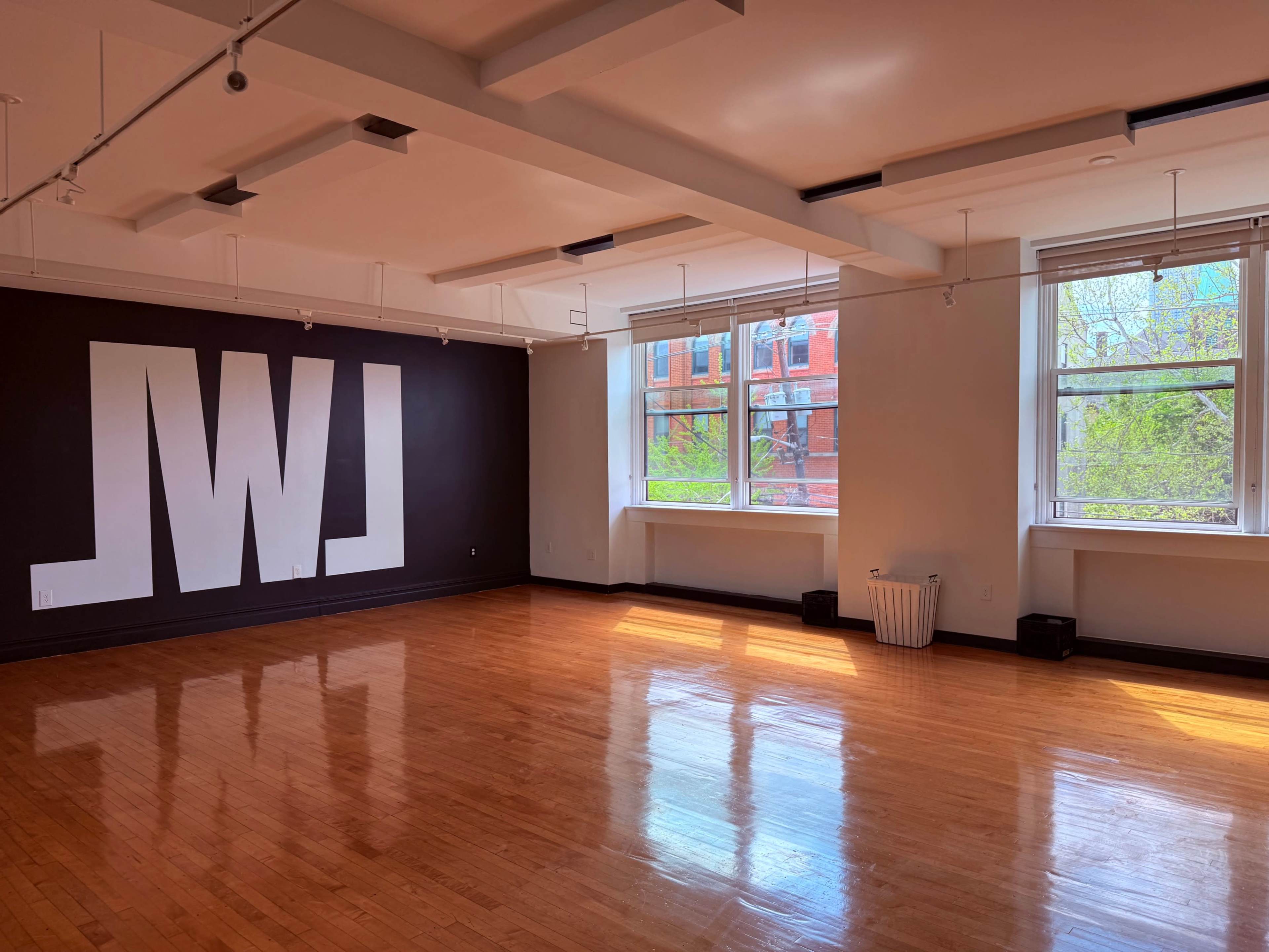 The image shows a spacious, well-lit studio with polished wooden floors and large windows, featuring a large white lettered mural on the black wall.