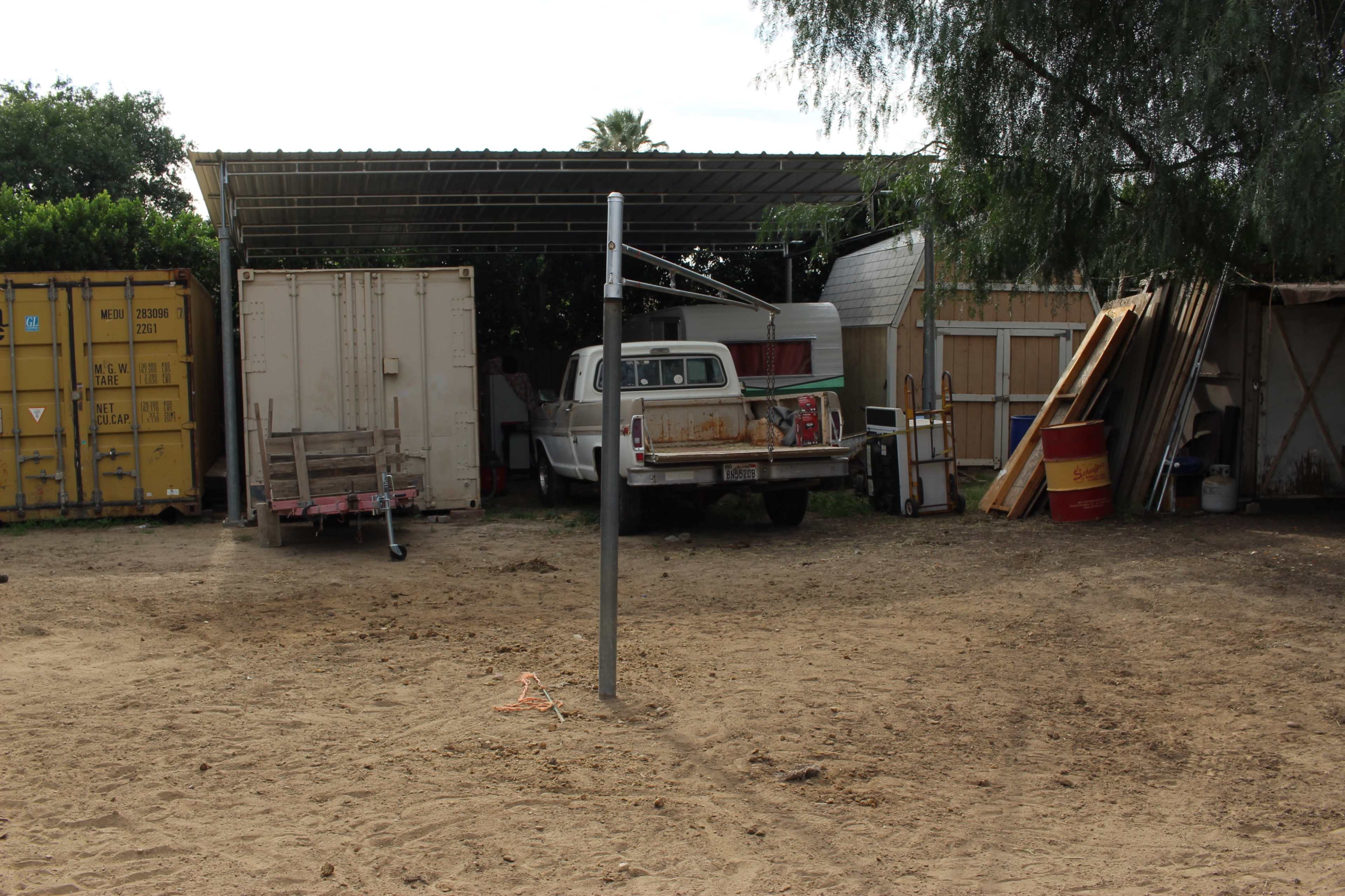 The image shows a dusty, open yard with a parked old pickup truck and various storage containers and items under a metal roof.