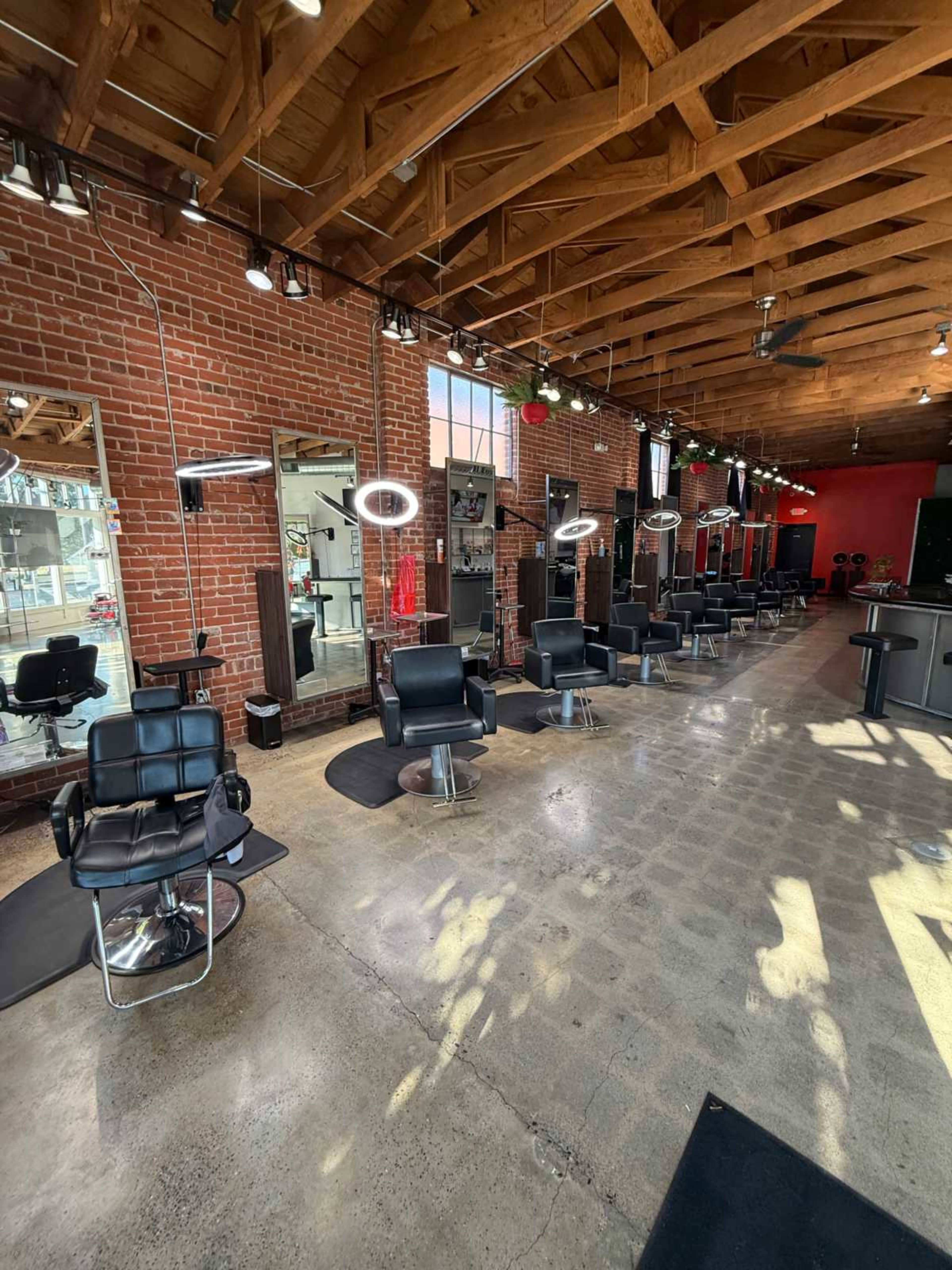 The image shows a modern hair salon featuring several styling chairs with mirrors, bright lighting fixtures, and a spacious layout with exposed brick walls.